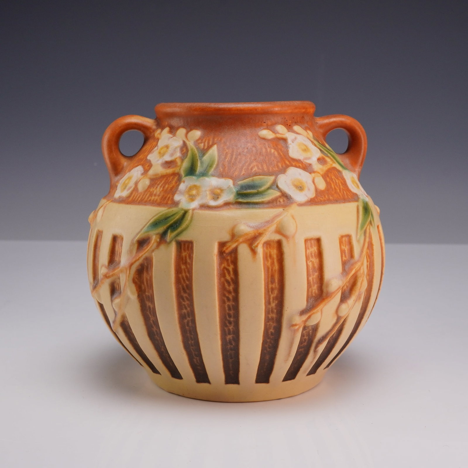 Roseville Pottery Cherry Blossom Two-Handled Vase: This art pottery vase is manufactured by Roseville Pottery Company of Zanesville, Ohio, in the Cherry Blossom pattern, which was introduced in 1933. The earthenware vessel features a globular body wit