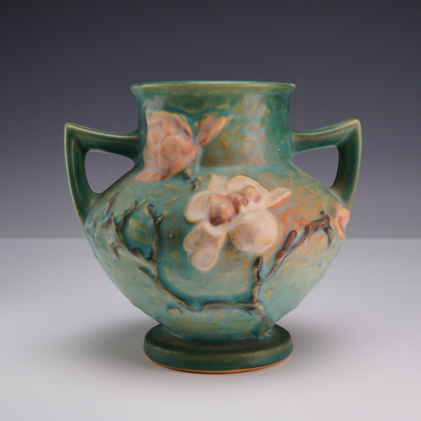 Roseville Blue Magnolia Pottery Double-Handled Vase 180-6: This Roseville Pottery double-handled vase is decorated in the Magnolia pattern, which was introduced by the company in 1943. The earthenware vessel features a squat, bulbous form with a flared rim an