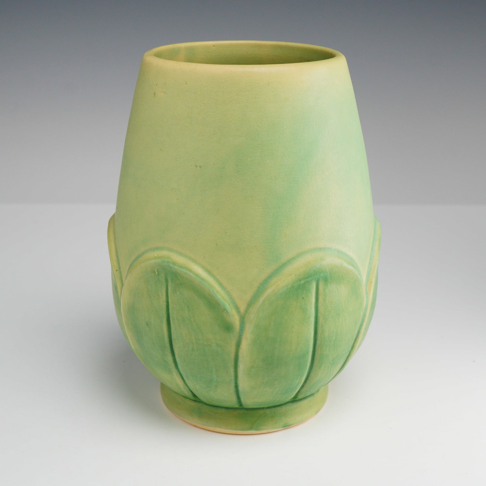 Weller Pottery Arts and Crafts Matte Green Leaf Vase: This earthenware pottery vase is manufactured by Weller Pottery of Zanesville, Ohio. The vessel features a tapered ovoid form supported by a slightly flared circular foot. The lower portion of the bod