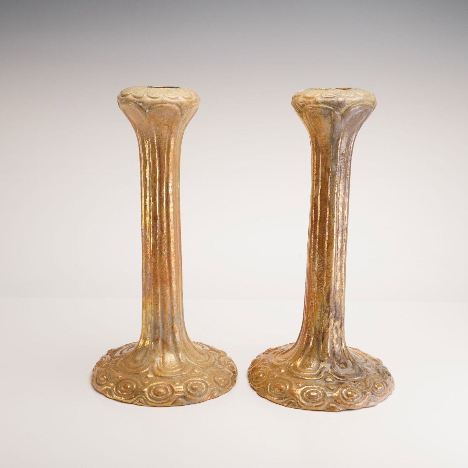 Pair of Leon Volkmar for Durant Kilns French Art Nouveau Iridescent Luster Ceramic Candlesticks: Each candlestick is composed of a slender, vertically fluted stem that flares at the top into a lobed nozzle and expands at the base into a wide, circular foot. The bases are decorated with relief-mol