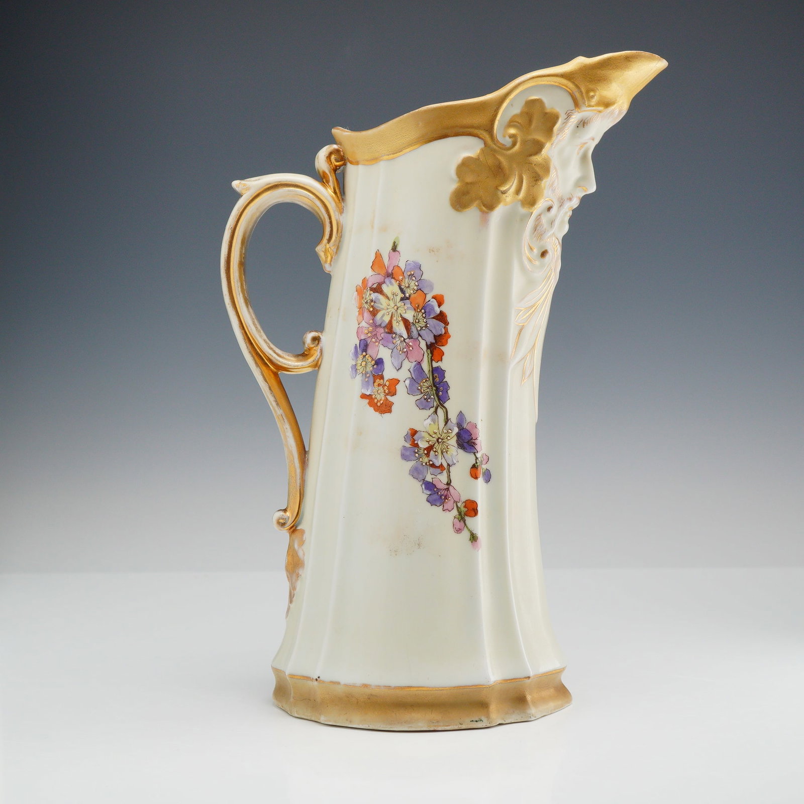 Victoria Carlsbad Austria Porcelain Pitcher with Floral and Face Accent: This is a tall porcelain pitcher produced by Victoria Carlsbad (Schmidt and Co.) in Altrohlau, Austria. The vessel features a faceted body and a spout molded into a high-relief mask of a bearded male 