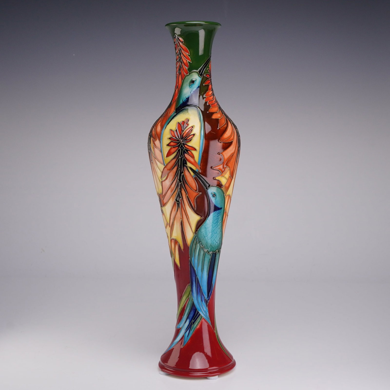 Moorcroft Pottery by Philip Gibson Limited Edition Vase: This glazed earthenware vessel features a complex tube-lined design depicting blue and teal hummingbirds amidst vibrant orange and yellow aloe blossoms. The background glaze transitions from a deep fo