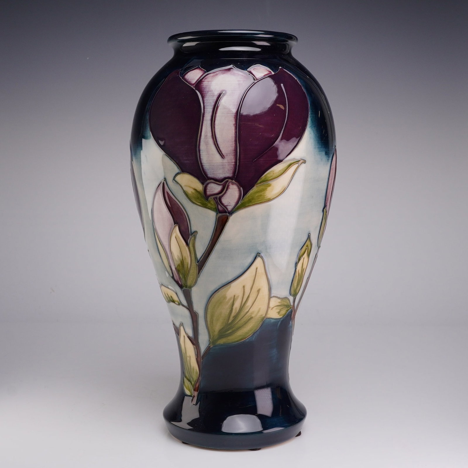 Moorcroft Pottery Baluster Vase, Magnolia Pattern: This ceramic earthenware vase features the Magnolia pattern produced by Moorcroft. The baluster shaped vessel is decorated with stylized magnolia blossoms in shades of deep purple and cream with green