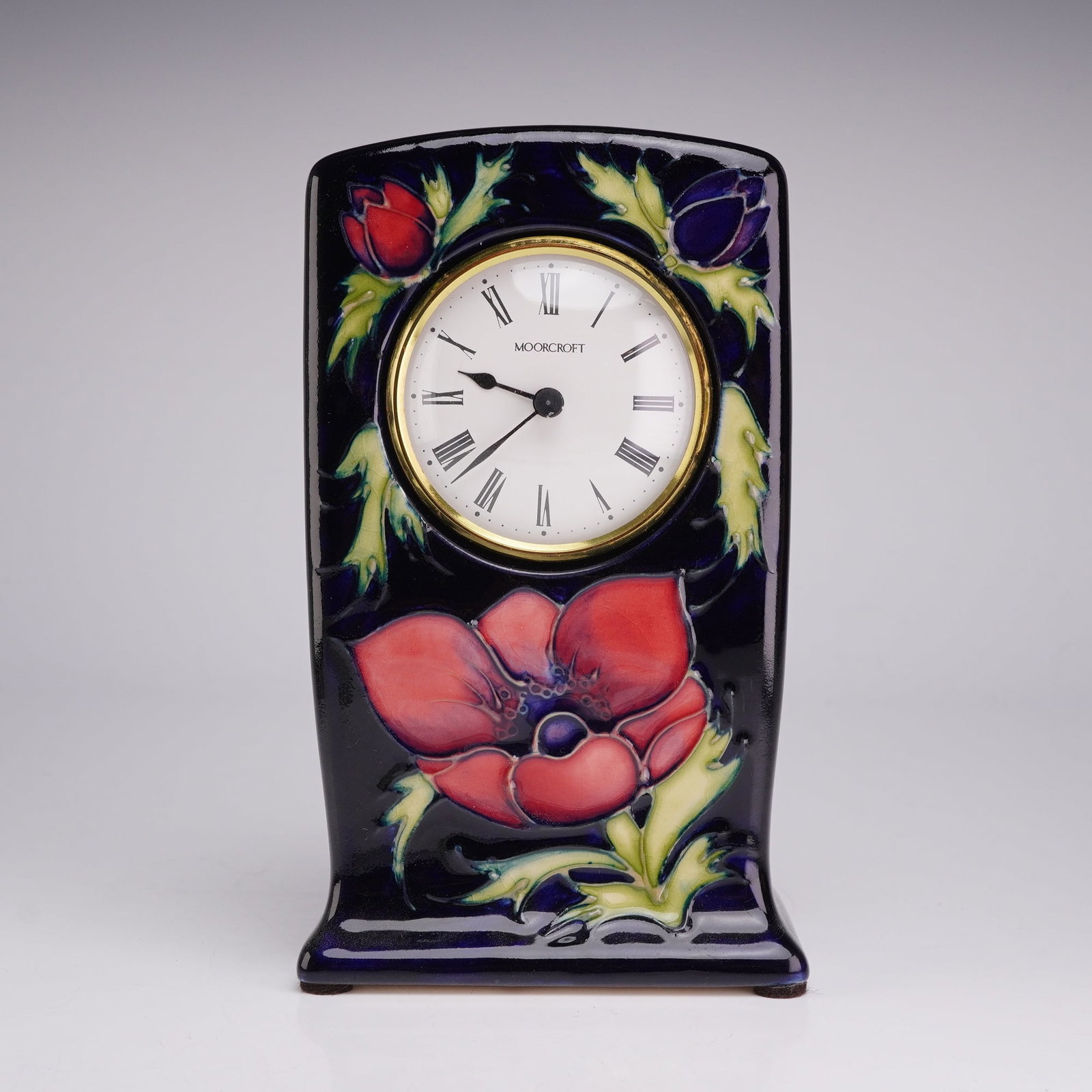 Moorcroft Pottery Table Clock, Anemone: The case features a rectangular, slightly tapered form with an arched top and a flared base. The decoration is applied using the tube-lining technique and depicts a large red anemone blossom at the bo