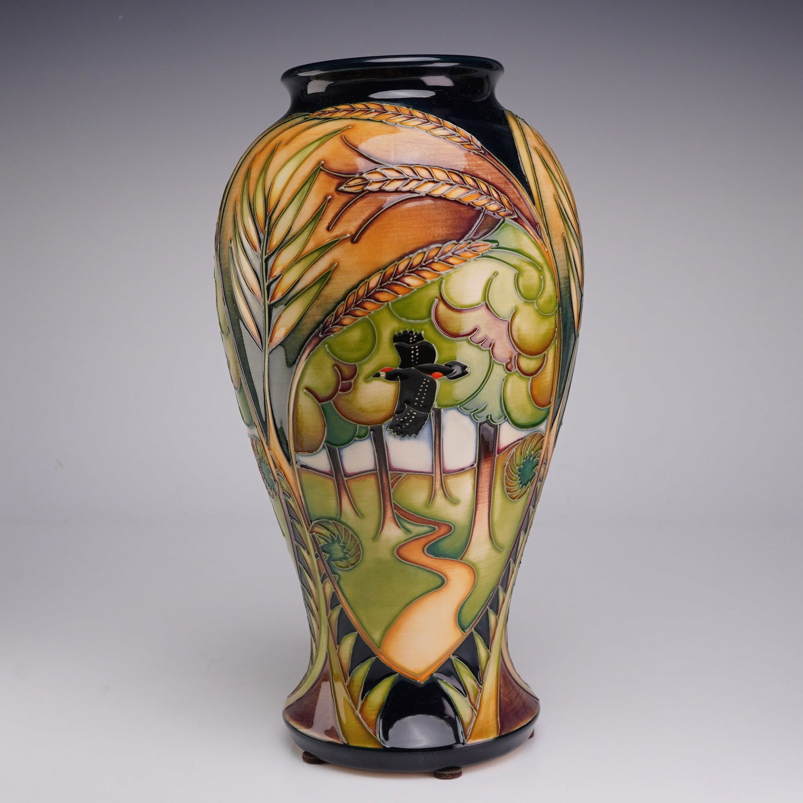 Moorcroft Pottery Landscape Medley Series Trial Vase, Woodland: The design depicts a stylized forest landscape with a winding path leading toward a white horizon, accented by a black and red woodpecker in flight. The central scene is framed by arching stalks of