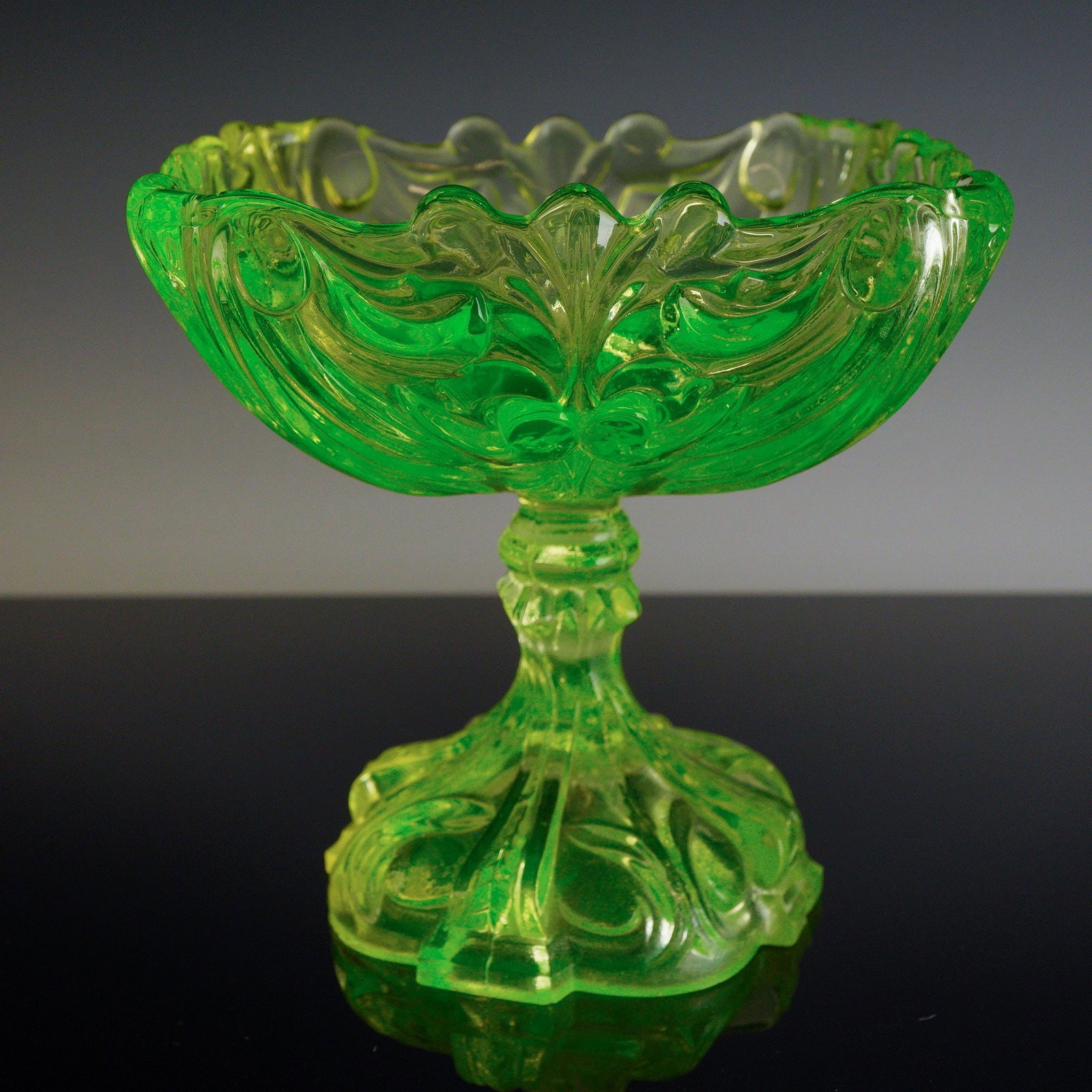 Victorian Style Ornate Pressed Uranium Glass Dish: This ornate standing glass dish is constructed from yellow-green pressed uranium glass, which fluoresces bright green under ultraviolet light. The piece features a wide bowl with a prominent scalloped