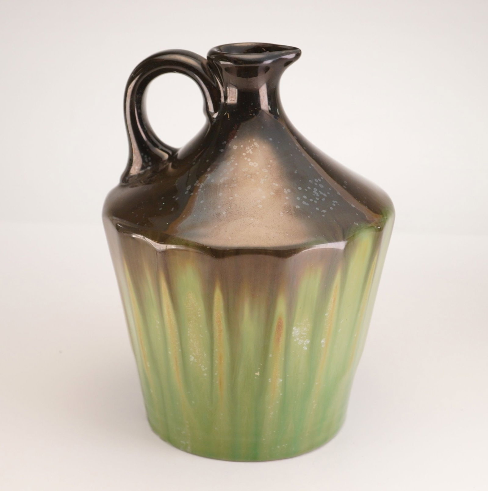 Fulper Pottery Green and Black Luster Musical Jug: Stoneware jug finished in a lustrous green and black glaze, featuring a short neck, integrated spout, and loop handle. The underside houses a wind-up musical mechanism. Label to base reads "Prague: Sa
