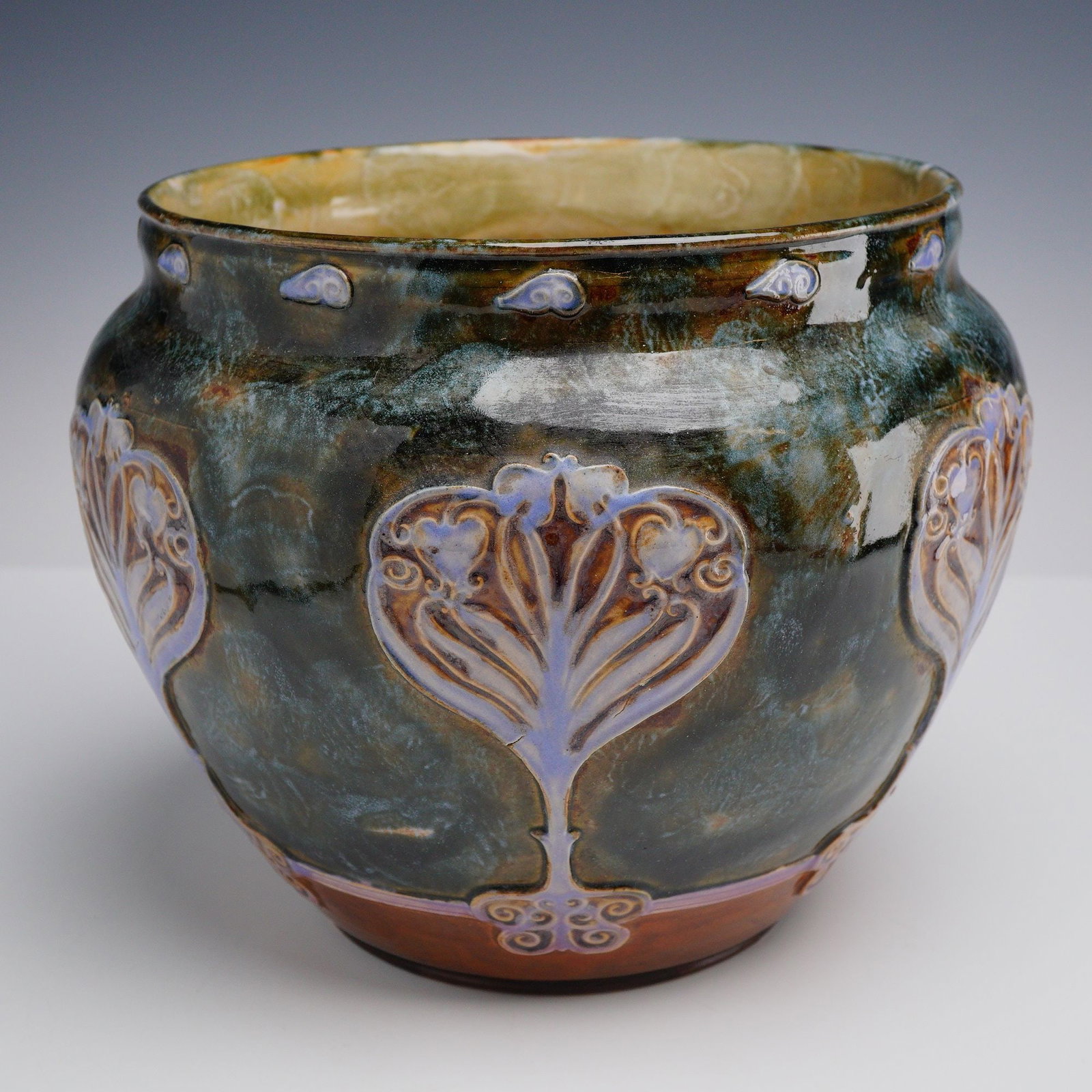 Doulton Lambeth Art Nouveau Jardiniere: This stoneware jardiniere features a squat, bulbous form with a wide, slightly flared mouth and a low center of gravity. The exterior is decorated with three relief-molded Art Nouveau foliate motifs, 