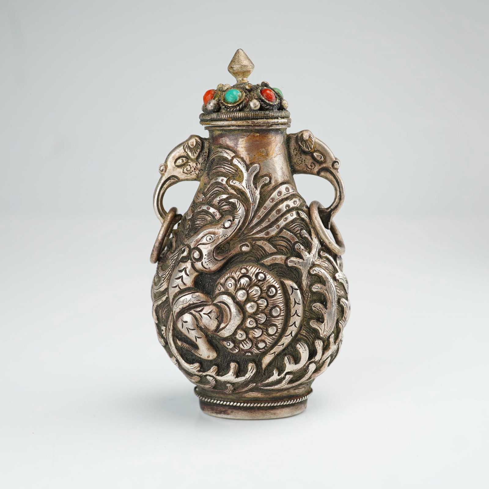 Mongolian Silver Repousse Snuff Bottle, Sea Creature and Waves: This Mongolian silver repousse snuff bottle with stopper features bird head handles, a sea creature amid a wave-like design, and a lotus flower accented with turquoise, coral, and lapis lazuli, with a