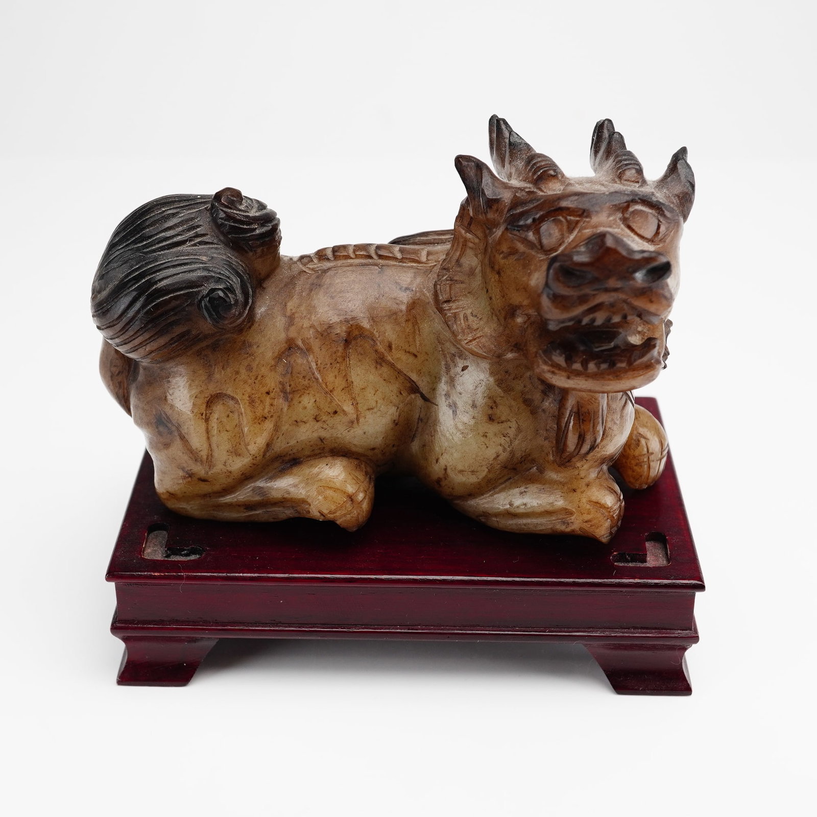 Chinese Carved Stone Foo Dog on Wooden Base: A Chinese carved hardstone figure of a mythical beast, or Foo dog, rendered in an archaic style. The creature is shown in a recumbent pose with a fierce expression, bulging eyes, an open mouth, and a 