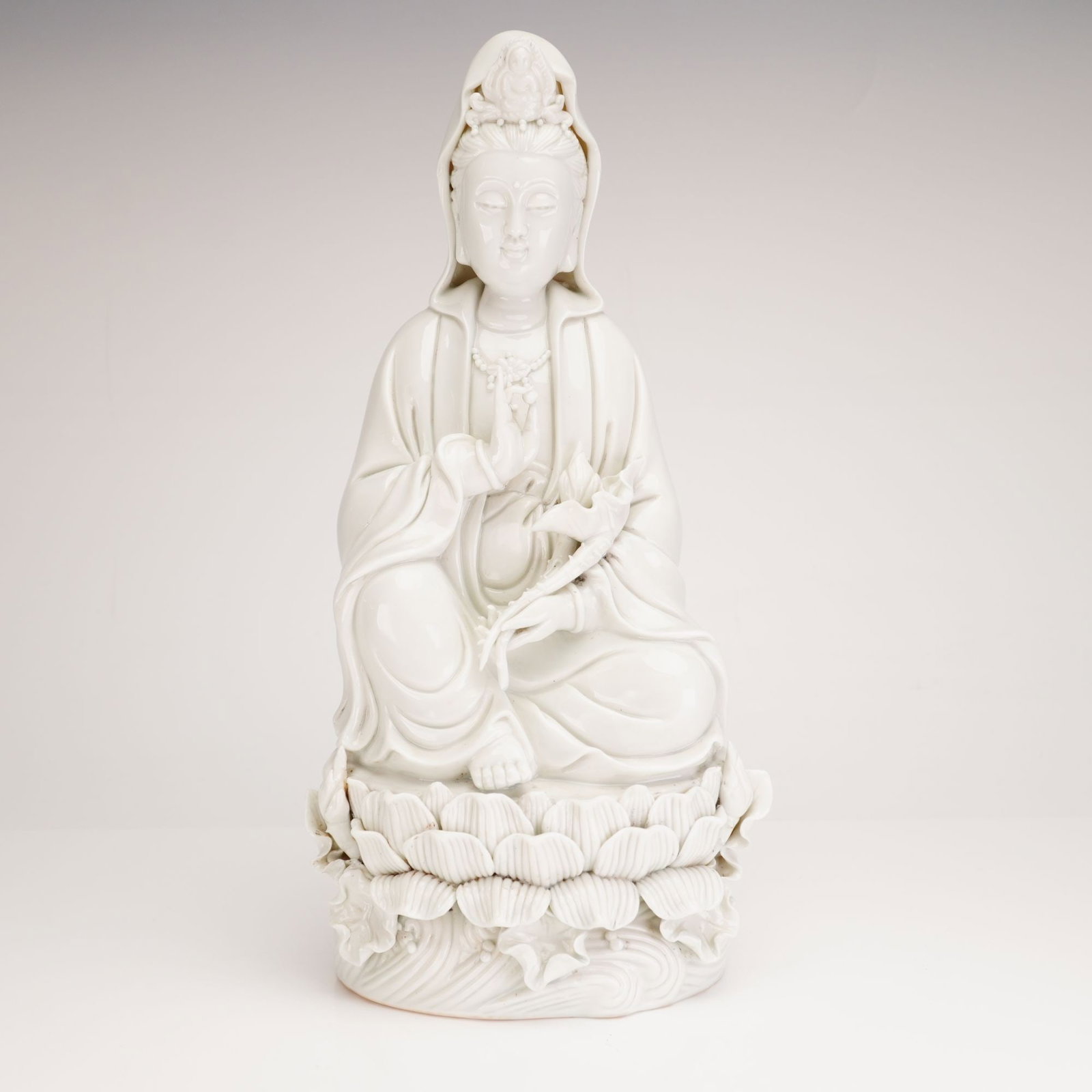 Chinese Dehua Style Blanc de Chine Porcelain Guanyin Figurine: A Chinese blanc de chine porcelain figurine of Guanyin, the Bodhisattva of Compassion. The figure is modeled seated in dhyanasana on a double-lotus pedestal above a base of stylized scrolling waves. S
