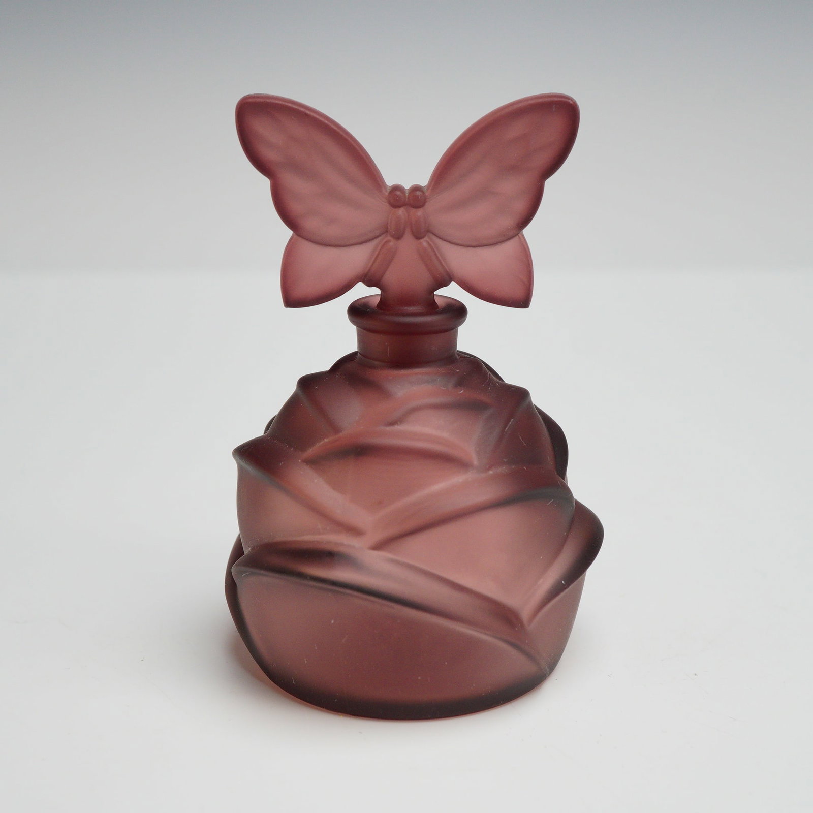 French Art Deco Frosted Glass Rosebud Perfume Bottle with Butterfly Stopper: This French art deco perfume bottle is crafted from smoky mauve glass with a frosted, satin finish. The body of the vessel is mold-blown in high relief to depict overlapping petals, forming a rosebud 