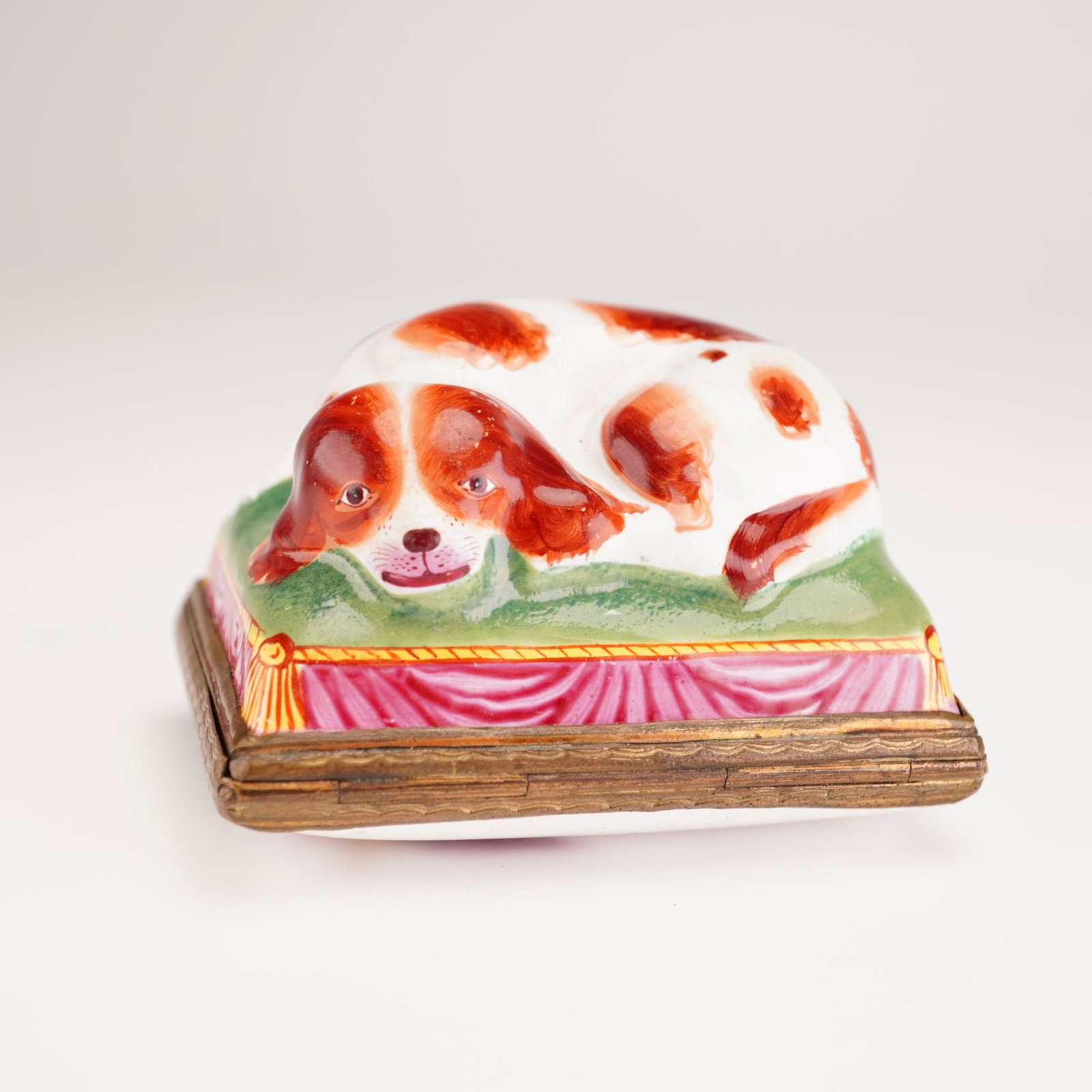 Antique English Enameled Bonbonniere, Cavalier King Charles Spaniel: This porcelain hand-painted treasure box is modeled in the form of a reclining Cavalier King Charles spaniel with iron-red markings, resting atop a green-tasseled cushion with pink-draped sides. The p