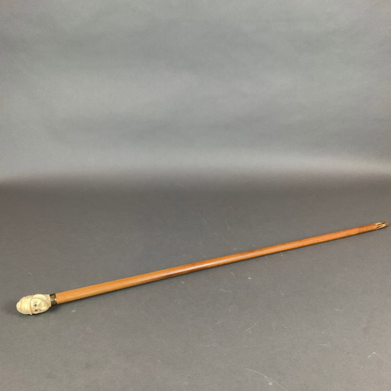 Wood Cane with Carved Bone Monkey Head: This antique walking stick consists of a tapered wood shaft topped with a handle intricately carved from bone. The handle is shaped as a monkey head with a hat on the handle and includes two inset