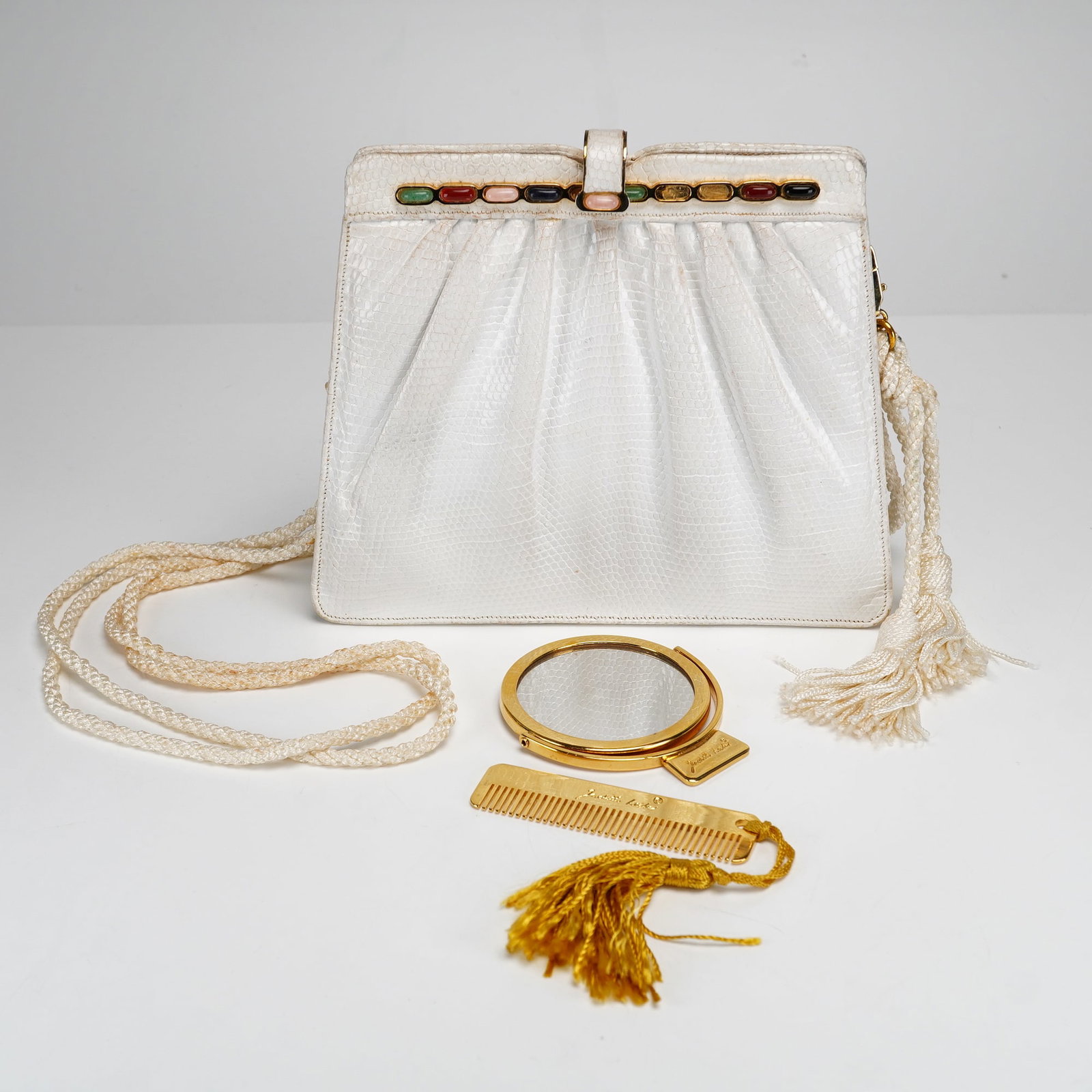 Vintage Judith Leiber White Lizard Skin Evening Bag: A white lizard skin shoulder bag with a gathered body and a cream-colored braided rope strap ending in a large tassel. The gold-tone metal frame is inset with a row of multi-colored oval cabochons.