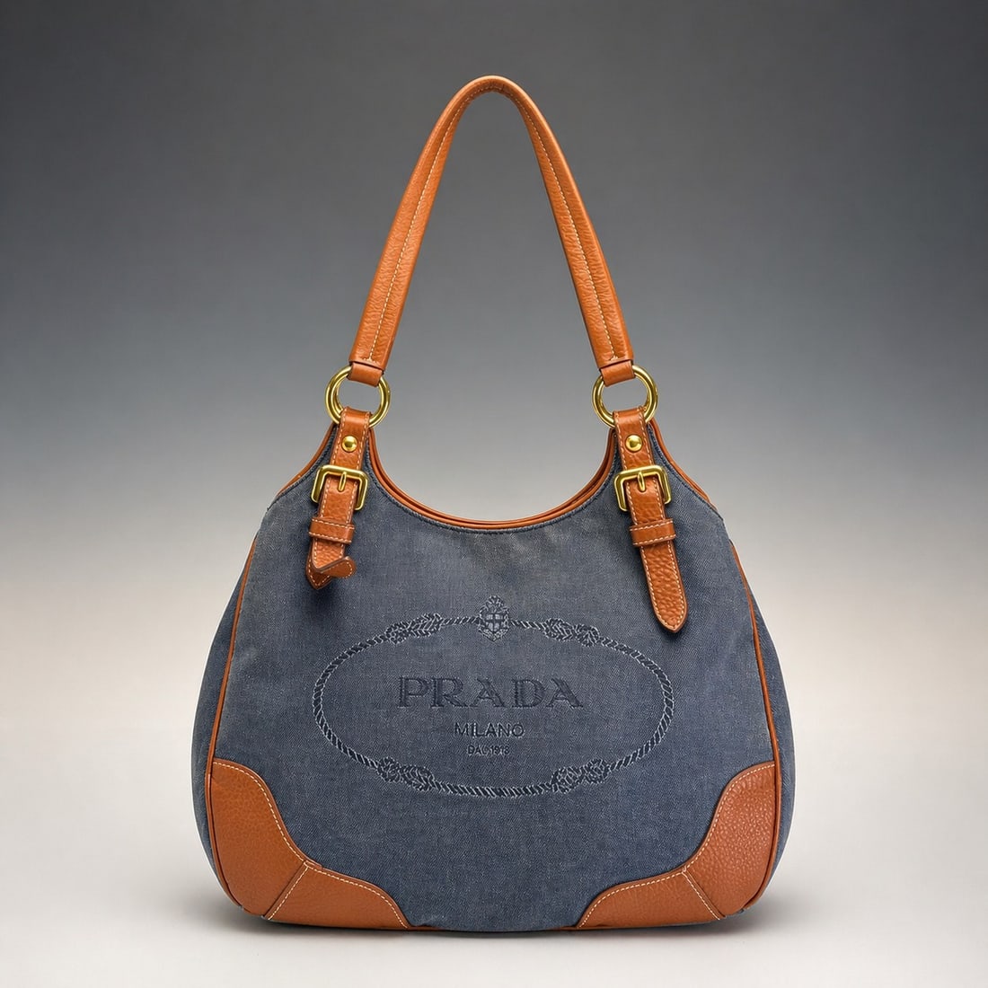 Prada Logo Jacquard Canvas and Leather Shoulder Bag: This Prada shoulder bag is constructed from blue denim-effect canvas with brown pebbled leather trim. The front of the bag features a large embroidered logo within an oval rope border, including the t