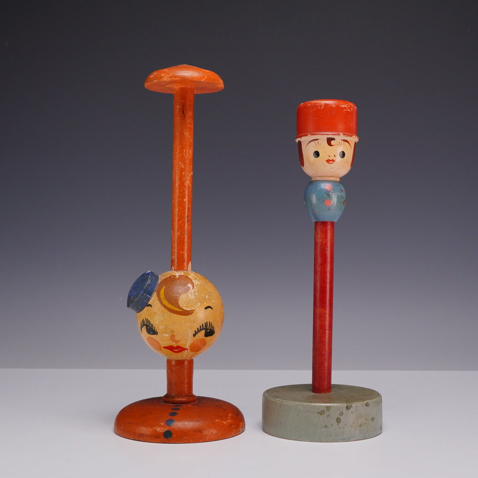Two Vintage Painted Wooden Novelty Hat Stands: A pair of vintage painted wooden novelty hat stands or millinery displays featuring stylized character heads. The larger stand consists of a tall orange-painted stem topped with a flat disc, featuring