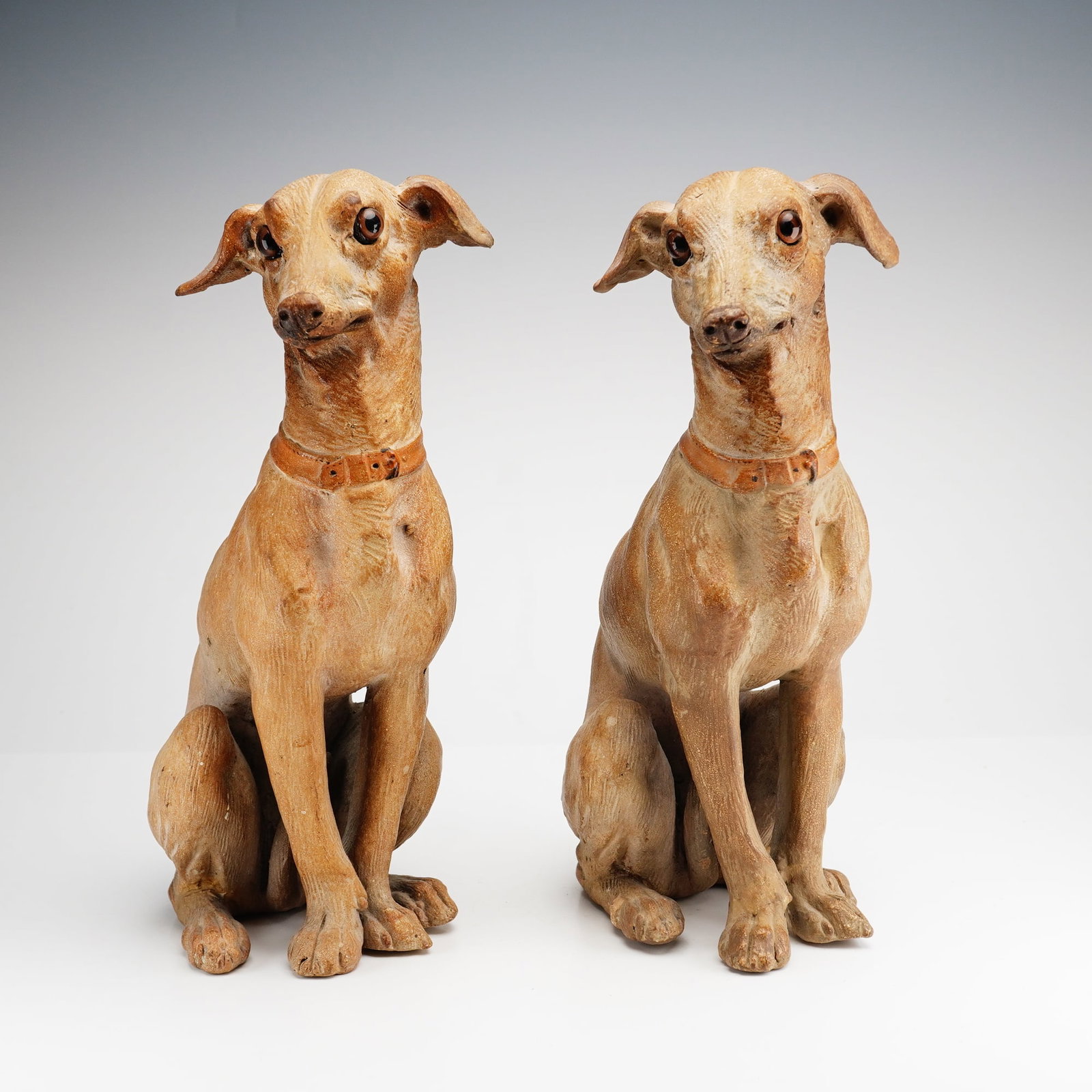 Pair of Italian Terracotta Greyhound Figural Statues: A pair of ceramic Italian Greyhound dog statues modeled in an alert, upright seated posture. Each figure features a mirrored turn of the head and is finished with a polychrome painted surface over ter