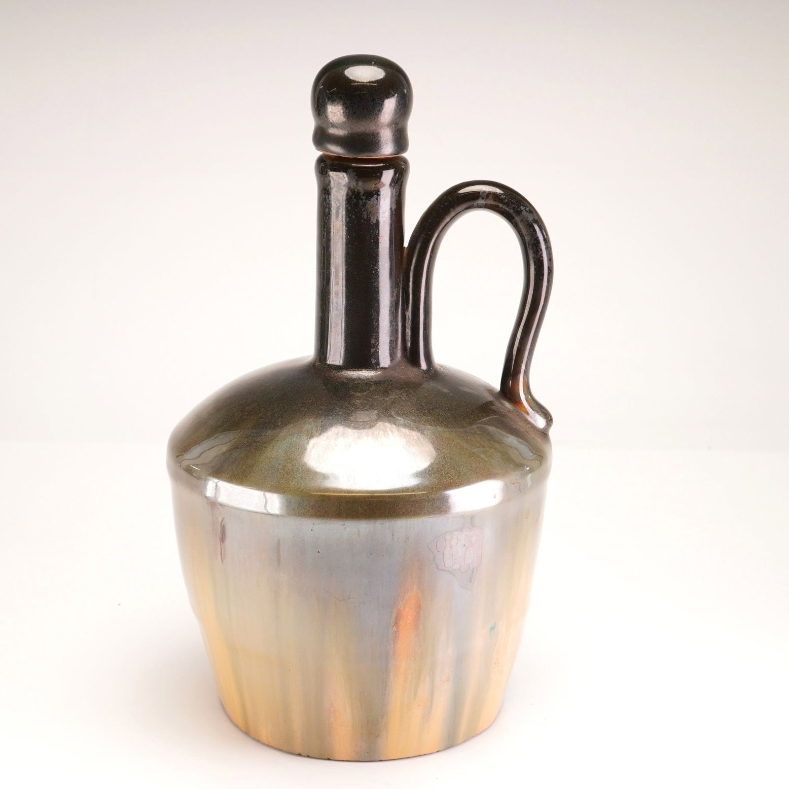 Fulper Pottery Silver and Black Luster Musical Jug: Stoneware jug finished in a lustrous black and silver glaze, featuring a long neck, spout, and applied handle. Accompanied by a round stopper. The underside houses a wind-up musical mechanism. Sticker