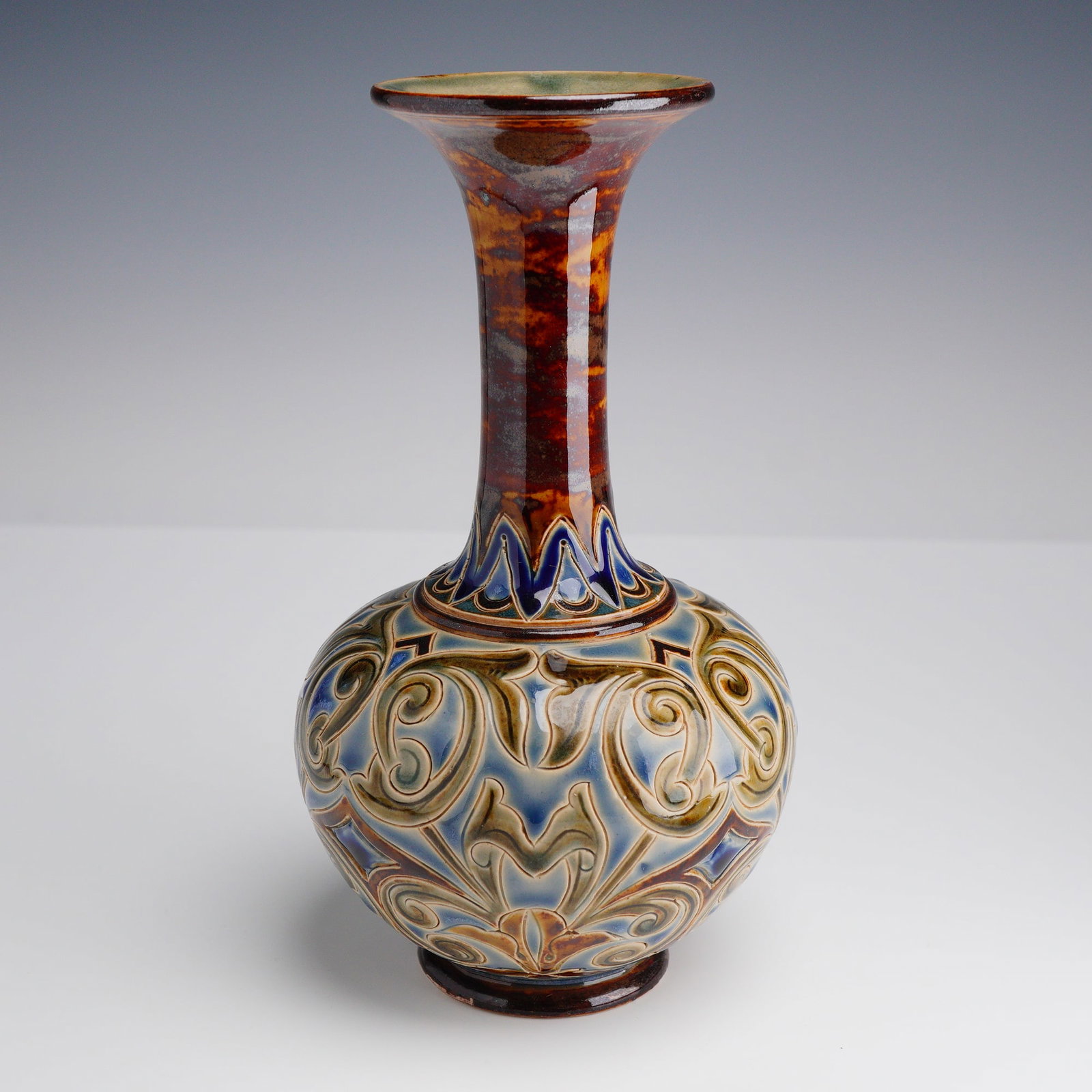 Doulton Lambeth Glazed Stoneware Vase: The vessel features a tall, tapering neck with a flared rim and a bulbous, spherical body. The body is decorated with hand-incised sgraffito scrolling foliate and stylized leaf patterns filled with hi