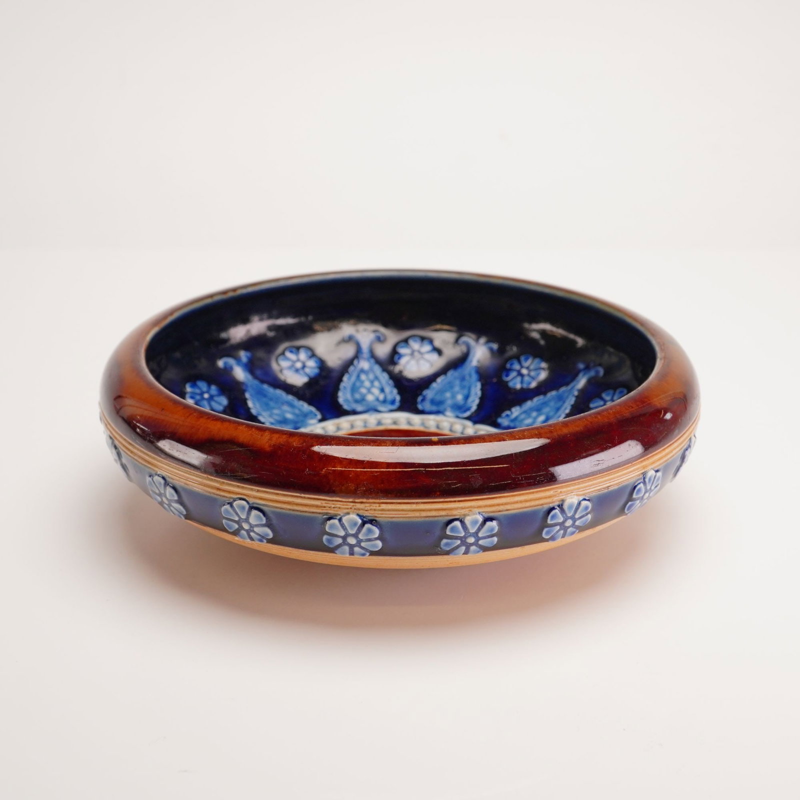 Doulton Lambeth Stoneware Bowl with Relief Decoration: This shallow circular vessel features a thick rounded rim and a low foot ring. The piece is decorated with a combination of molded and applied relief under a multi-tone glaze. The wide rim and undersi