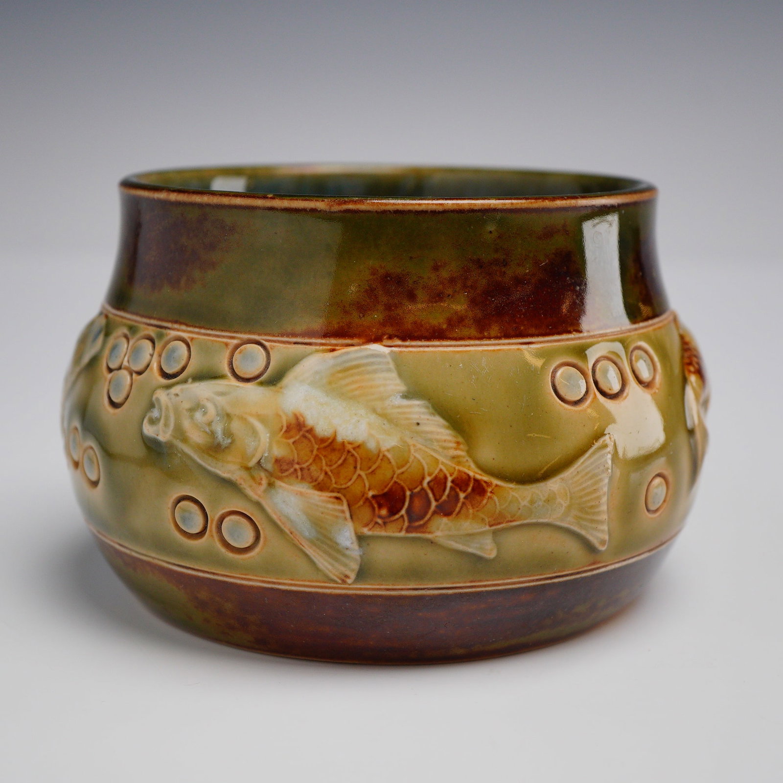 Royal Doulton Lambeth Stoneware Small Fish Pot: The vessel features a constricted neck and slightly flared rim. The exterior is decorated with relief-molded figures of fish, possibly carp or koi, swimming among raised circular bubbles against an ol