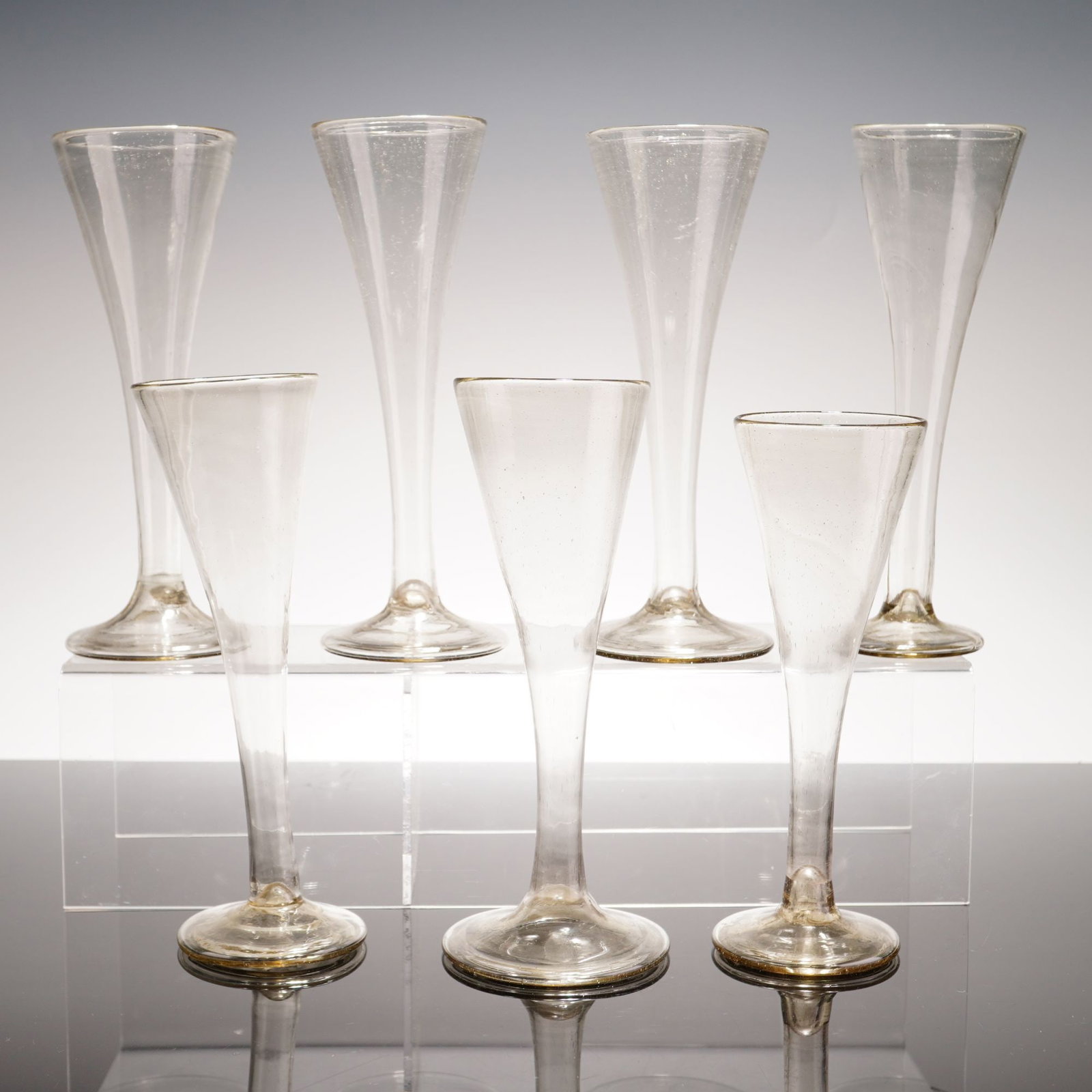 7pc Hand-Blown Hollow Stem Glass Champagne Flutes, c. 1800s: A set of seven handmade glass flutes featuring hollow stems and trumpet-shaped bowls. These vessels are hand-blown, which accounts for the observable variances in size, height, and bowl shape througho