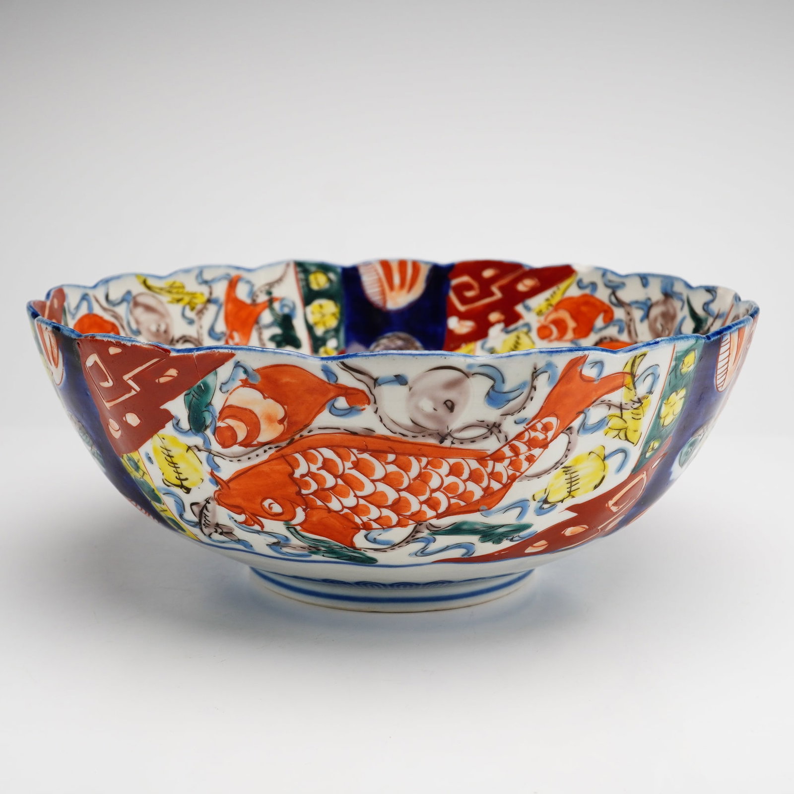 Japanese Imari Porcelain Scalloped Bowl with Carp Motif: A Japanese Imari porcelain bowl of deep circular form featuring a scalloped or foliate rim. The piece is decorated in a traditional palette using underglaze cobalt blue and overglaze iron-red, yellow,
