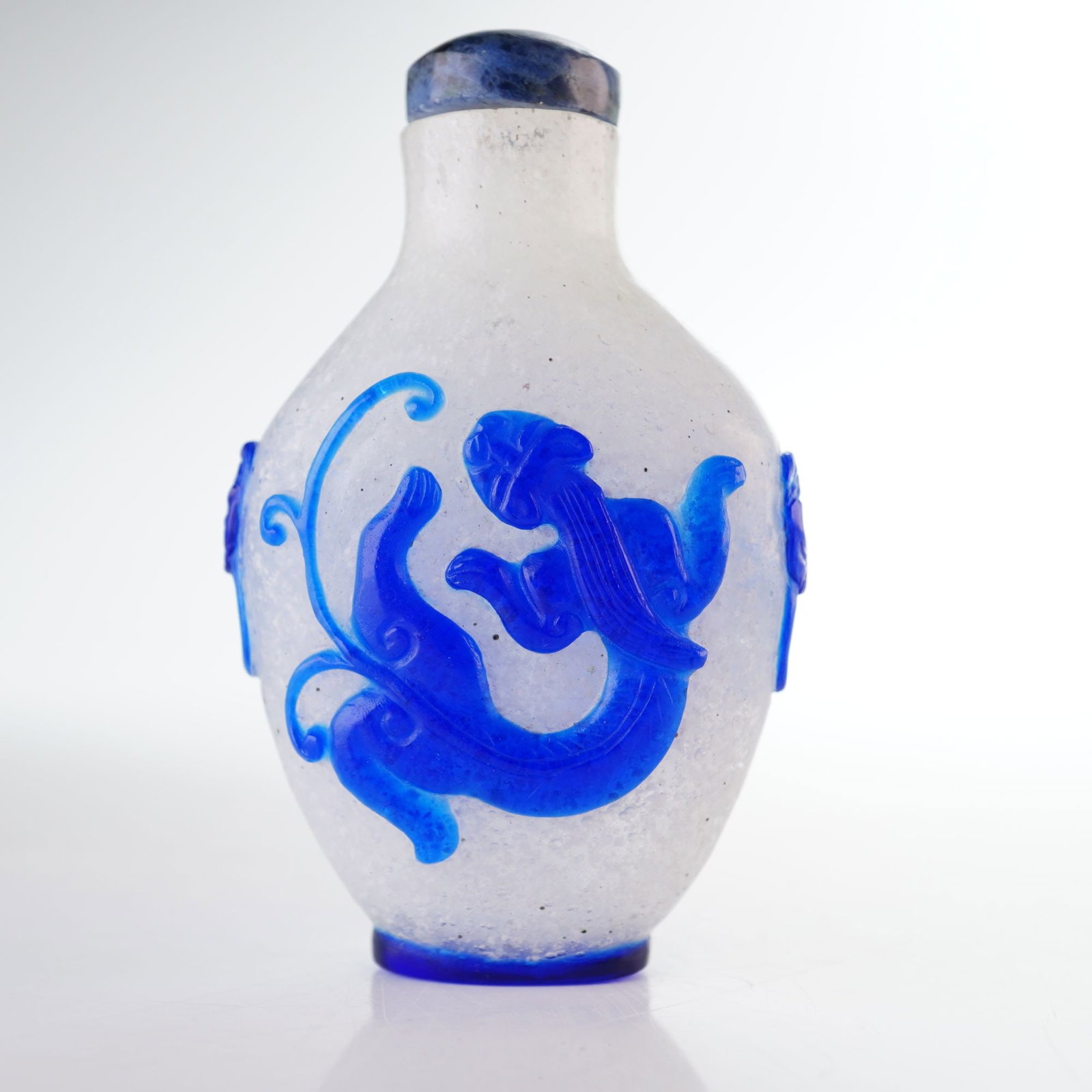 Peking Glass Snuff Bottle, Blue Overlay Chilongs and Faux Lion Handles: A clear Peking glass snuff bottle featuring a snowflake pattern adorned with high-relief blue overlay depicting Chilongs and simulated lion mask handles with rings. It is topped with a mottled dark bl