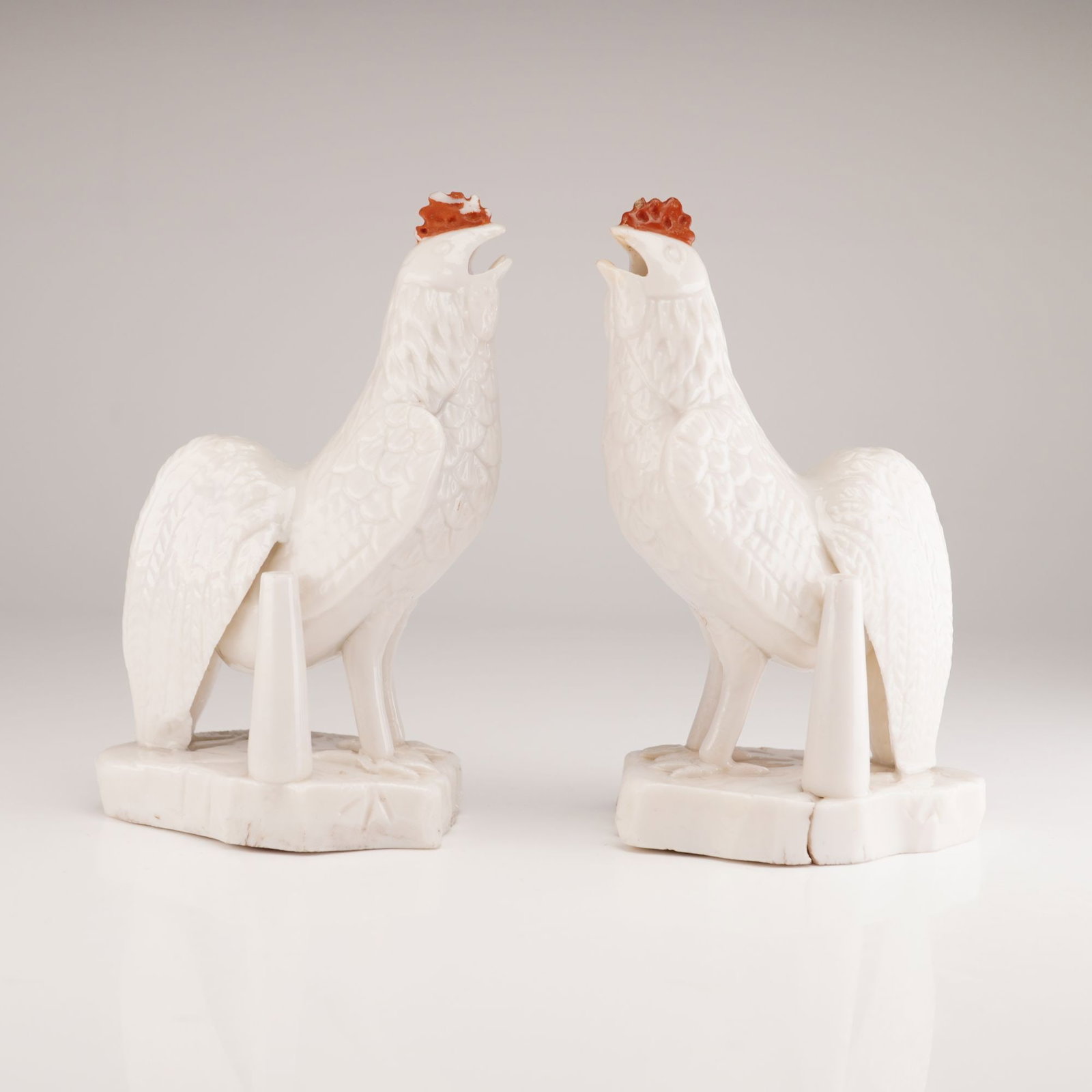 Pair of Chinese Blanc de Chine Porcelain Rooster Joss Stick Holders: A pair of porcelain figural joss stick holders in the style of Dehua blanc de chine, each modeled as a crowing rooster with head raised and beak open. The figurines are crafted from white glazed porce
