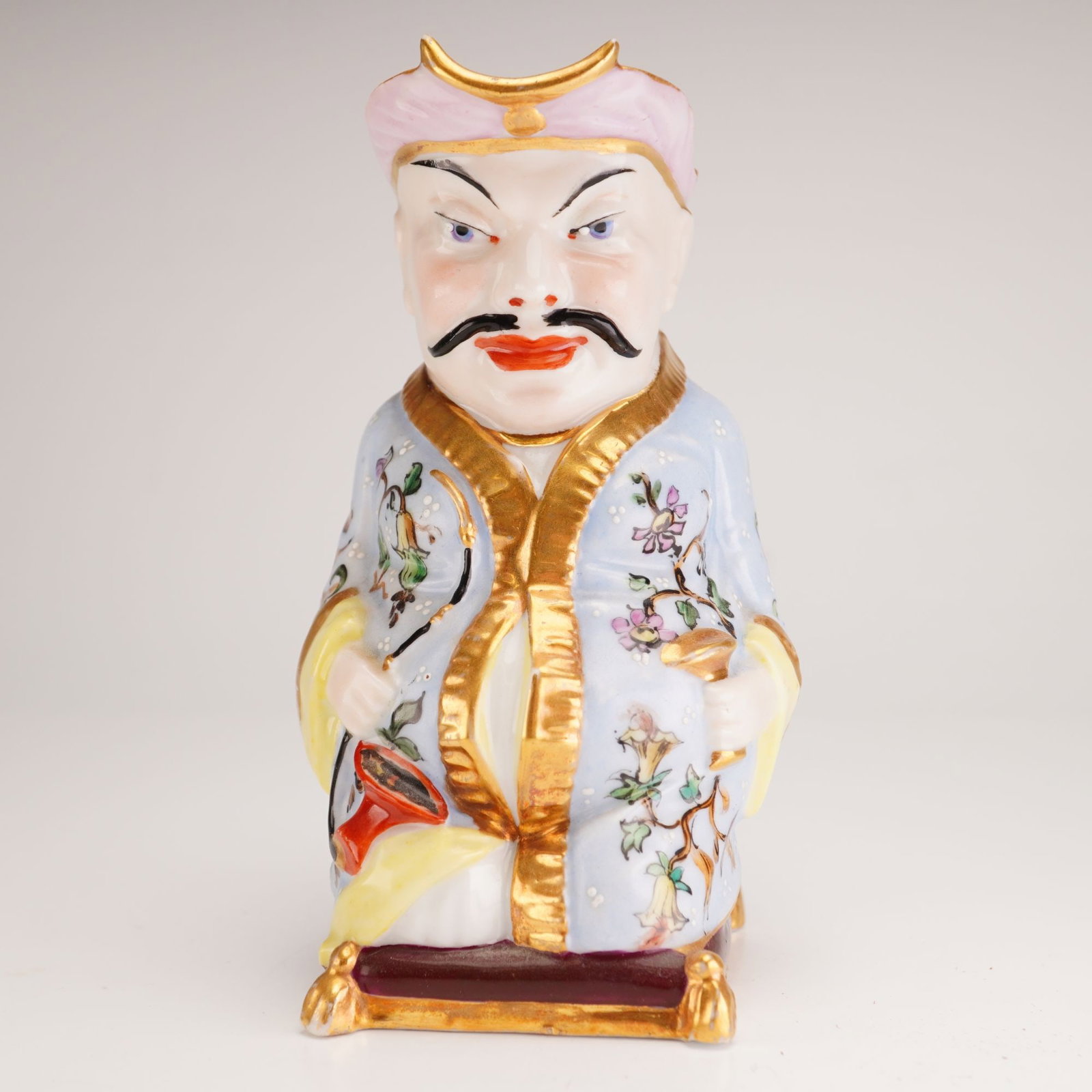 Ornate Chinoiserie Porcelain Cream Pitcher: This porcelain cream pitcher is modeled in the form of an ornate Chinese seated man, characterized by a stern expression, black mustache, and high-arched eyebrows. The figure is depicted in the Chinoi