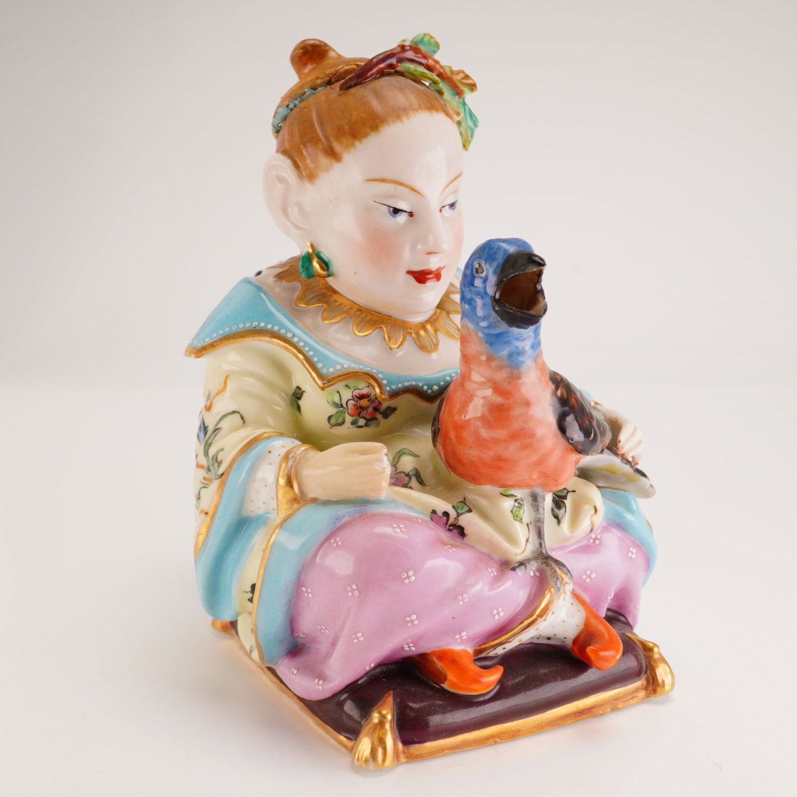 Porcelain Lidded Teapot in the Chinoiserie Style: A lidded porcelain figural teapot or vessel designed in the Chinoiserie style. The piece depicts a seated woman in stylized Eastern attire, featuring a pale yellow robe with floral enamel decoration, 