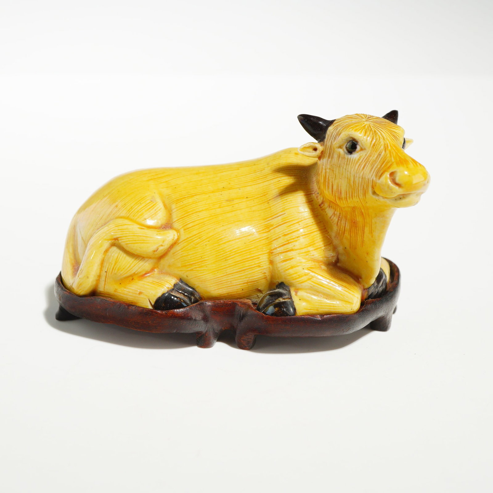 Chinese Ochre Glazed Ceramic Recumbent Ox Figurine with Wood Stand: A Chinese glazed ceramic figurine of a recumbent ox, also referred to as a water buffalo, depicted in a resting position with its head turned slightly to the right and its tail resting against its fla
