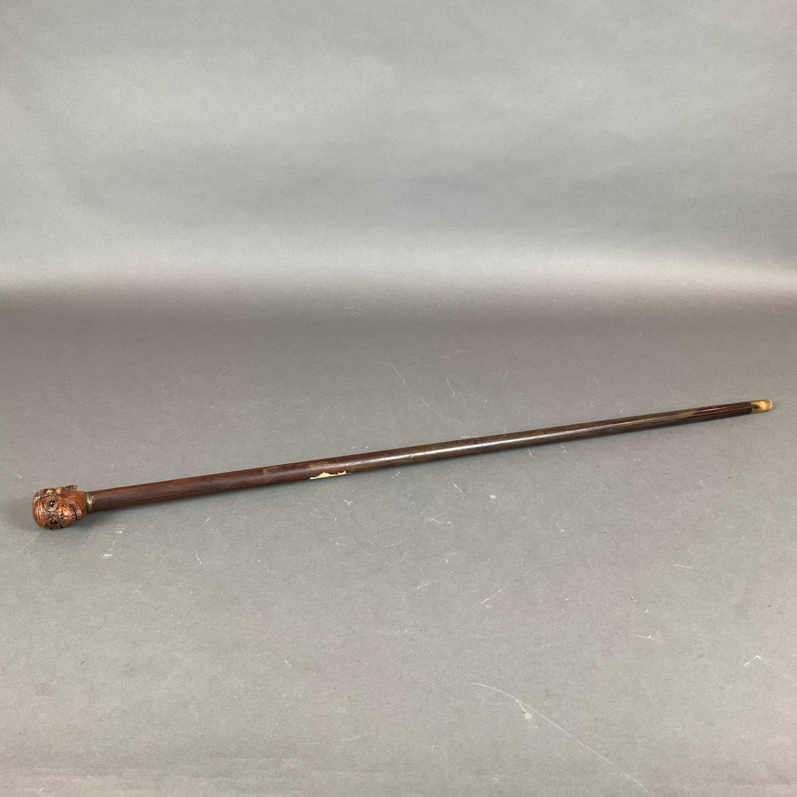 Carved Wood Monkey Head Walking Cane: This antique walking cane consists of a tapered wooden shaft topped with a hand-carved handle. The handle is fashioned into a monkey head with a braid, featuring detailed facial anatomy including a ba