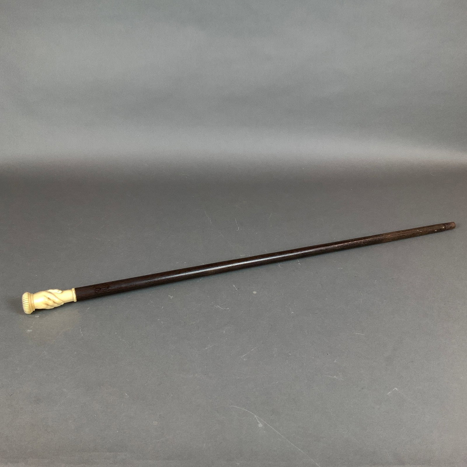 Wooden Walking Cane with Carved Bone Handle: This antique walking stick features a tapered shaft made of dark-stained wood paired with a decorative carved handle. The handle is crafted from bone and depicts a hand holding the cane, a traditional