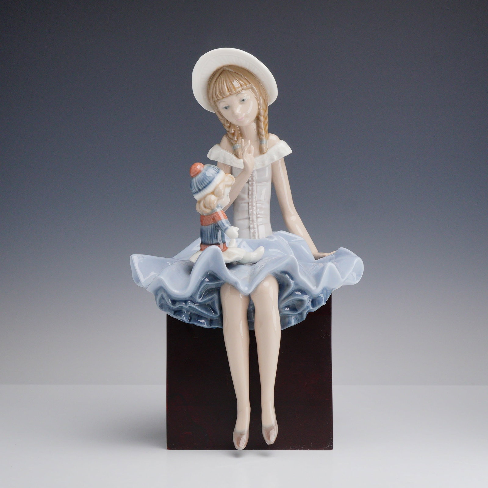 Lladro Porcelain Figurine Suzy and Her Doll Glazed 01001378: The figurine depicts a seated young girl with blonde braids, wearing a wide-brimmed white hat with a blue ribbon and a white off-the-shoulder bodice. She is dressed in a large, ruffled, light blue ski