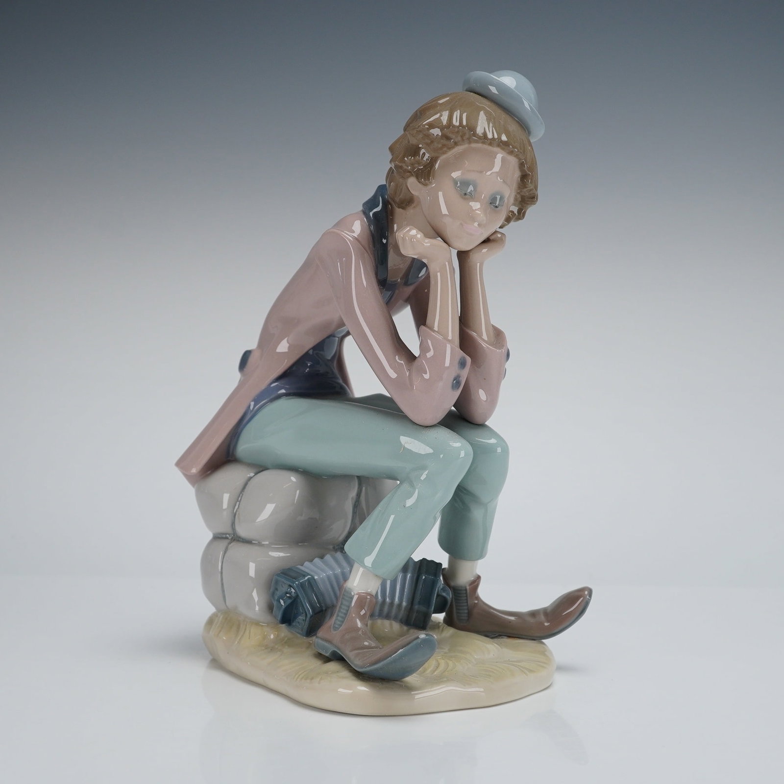 Lladro Porcelain Figurine, Thinking Clown (01005058): The sculpture depicts a seated male figure, styled as a melancholic Pierrot, resting his chin in his hands. The figure is seated atop stacked gray sacks or bales. He is dressed in pastel-colored cloth