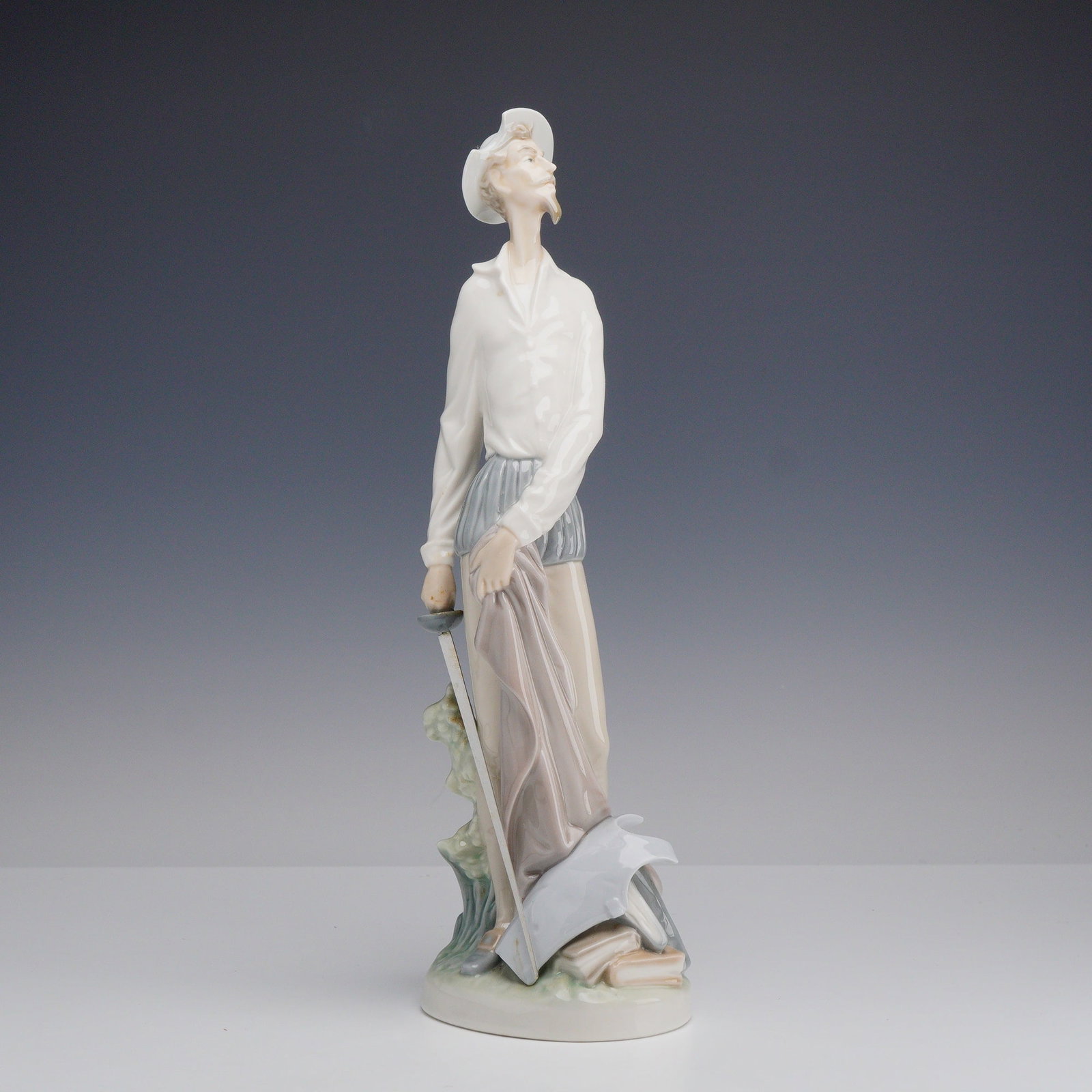 Lladro Porcelain Figurine, Don Quixote Standing 01004854: The figurine depicts the literary character in a slender, elongated form, posed looking upward. He is dressed in a white shirt and grey puffed breeches with a white wide-brimmed hat. He holds a separa