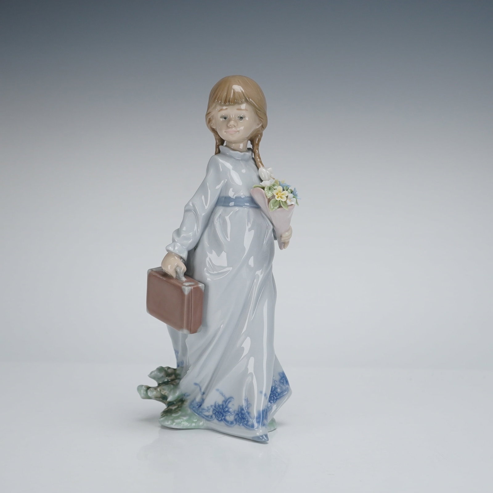 Lladro Porcelain Figurine School Days 7604: A Lladro porcelain figurine, titled "School Days," model number 7604. The glazed figurine depicts a young girl with brown pigtails, wearing a pale blue gown cinched at the waist, standing on a base de