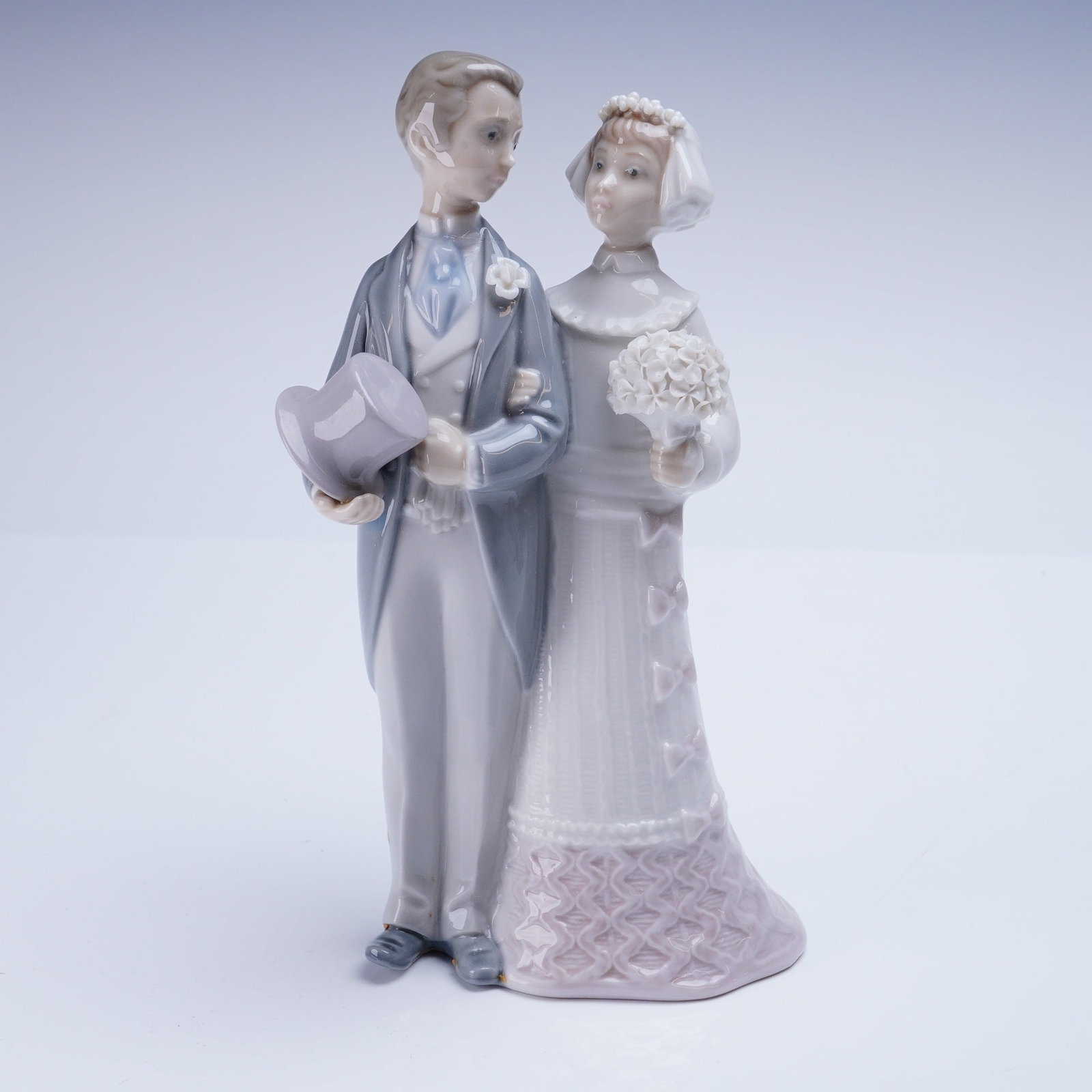 Lladro Porcelain Figurine Wedding 1004808: A single glazed Lladro Porcelain Figurine, titled Wedding, model number 1004808, designed by Julio Fernandez. The piece depicts a young bride and groom in formal wedding attire. The groom is dressed i