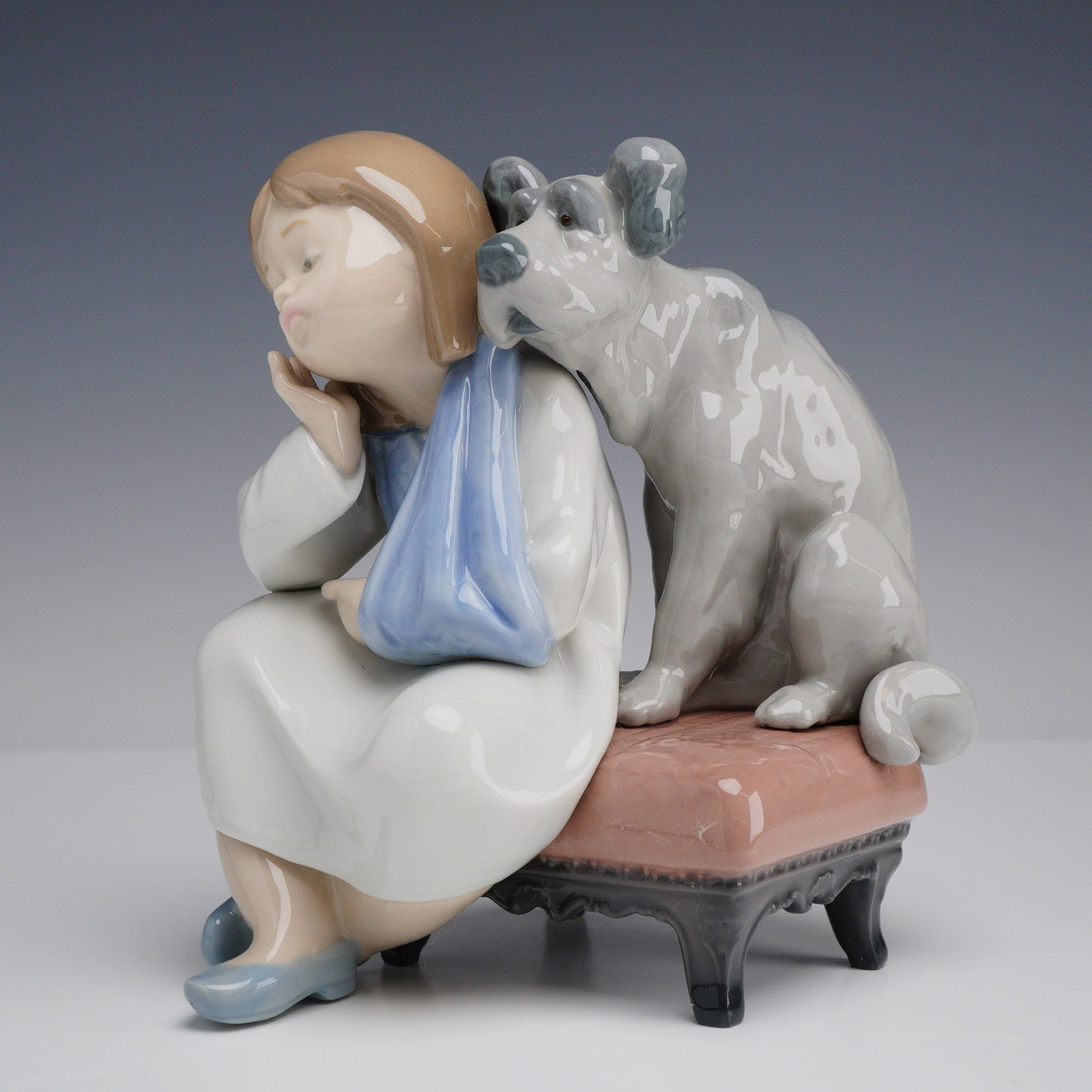 Lladro Porcelain Figurine We Can't Play 01005706: This Lladro glazed porcelain figurine depicts a young girl and her companion dog. The girl is modeled with short brown hair and her left arm in a blue sling, sitting in a pensive pose on a pink uphols