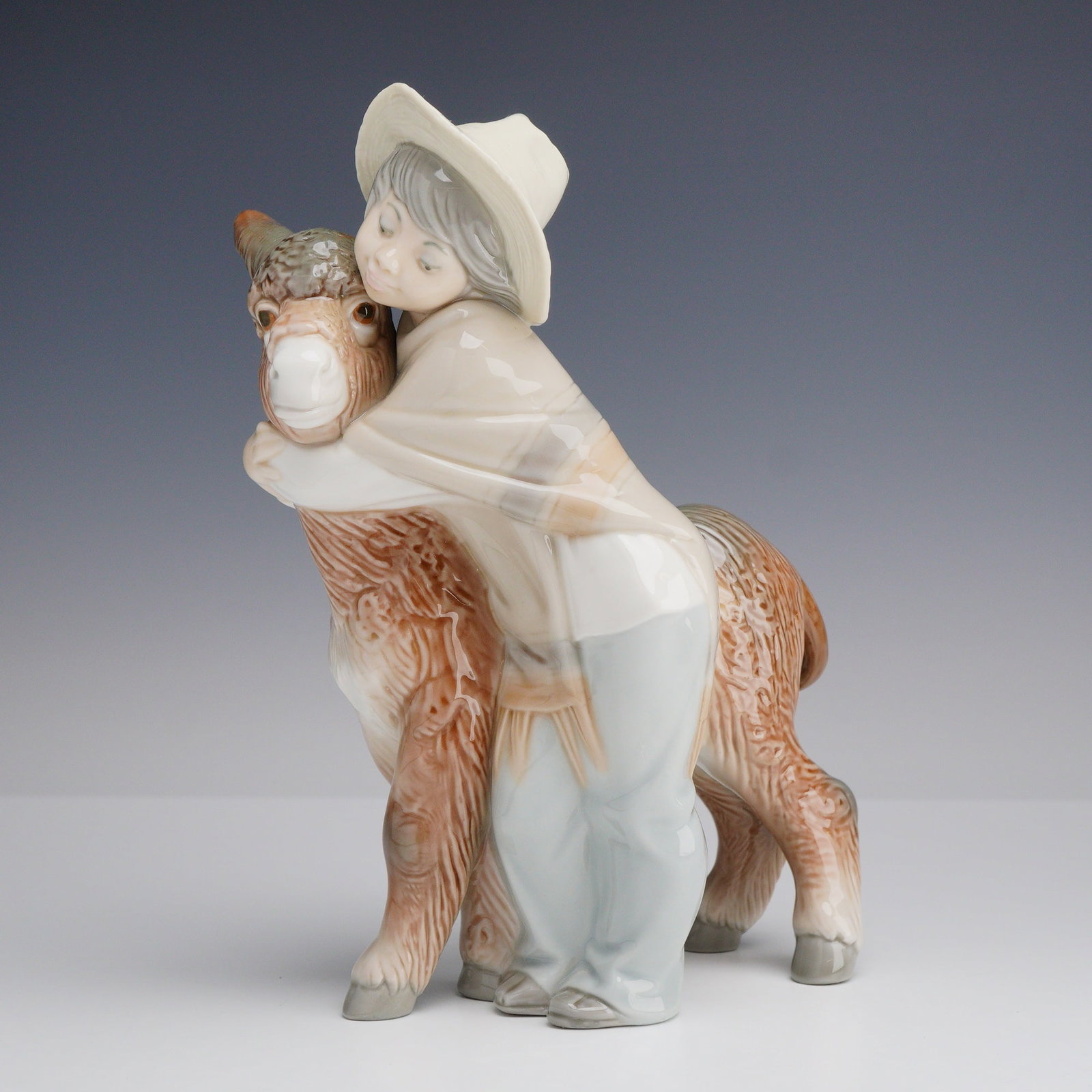 Lladro Porcelain Figurine Platero and Marcelino 01001181: This Lladro porcelain figurine depicts a young boy in a wide-brimmed hat and poncho affectionately hugging a small donkey. The piece is rendered in glossy glazed porcelain with a palette of soft brown