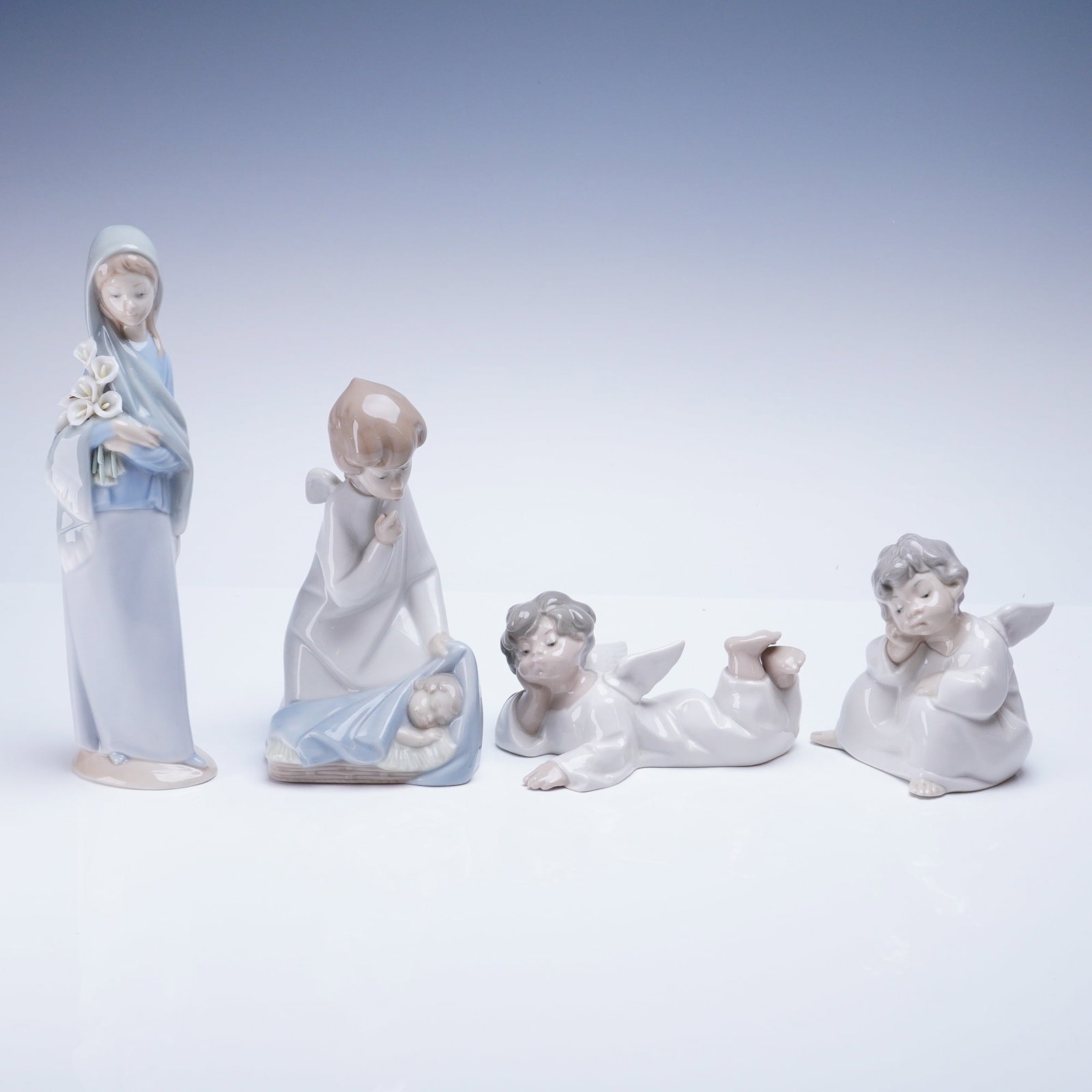 Collection of 4 Lladro Figurines Madonna with Angels: A collection of four glazed porcelain figurines manufactured by Lladro, Spain, depicting elements of a Nativity scene. The pieces are executed in the characteristic soft-focus style, utilizing a palet