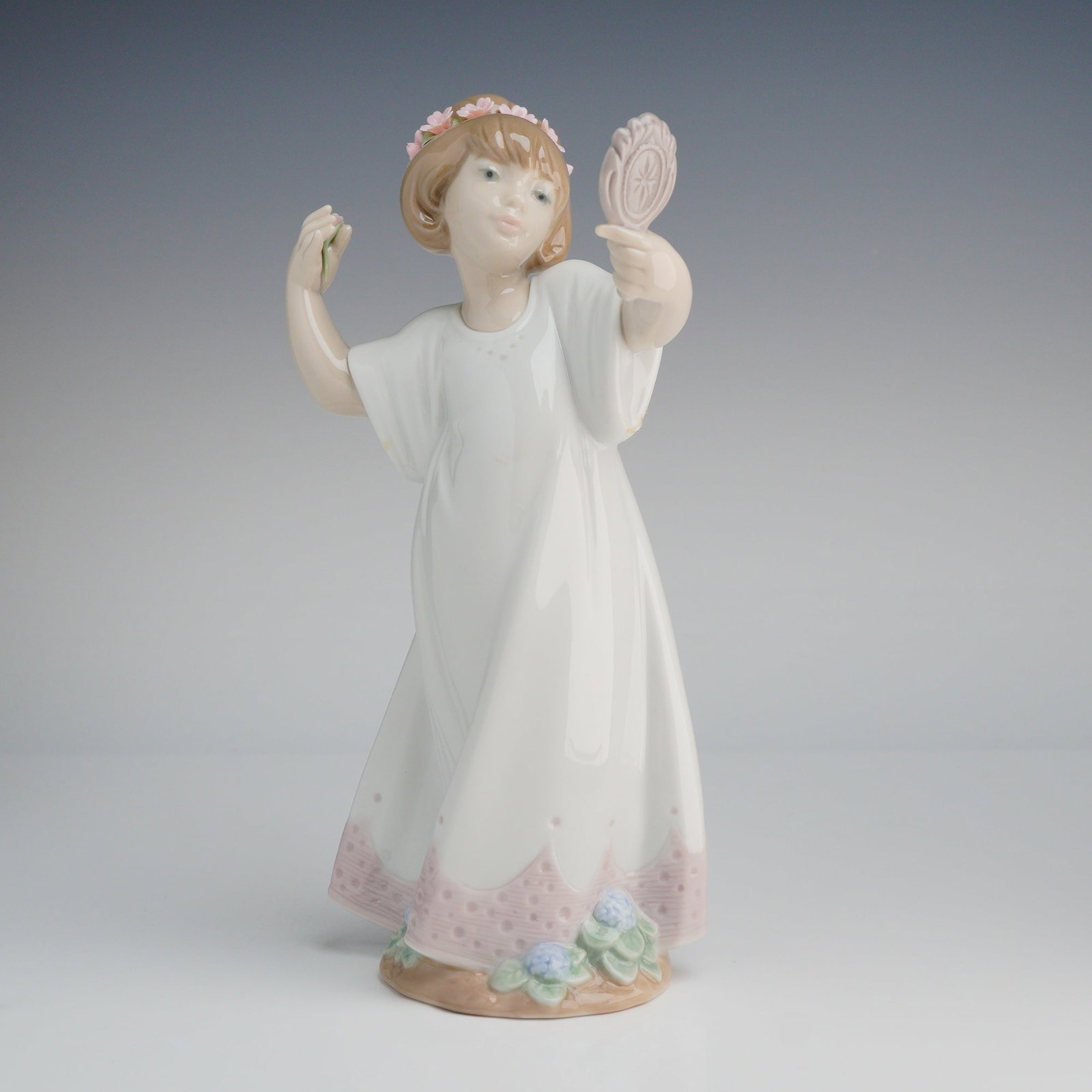 Lladro Porcelain Figurine Delightful Girl with Mirror 6806: A Lladro porcelain figurine, model number 6806, titled "Delightful." The figurine depicts a young girl standing in a long, flowing white glazed gown. The base of the gown features pink scalloped trim 