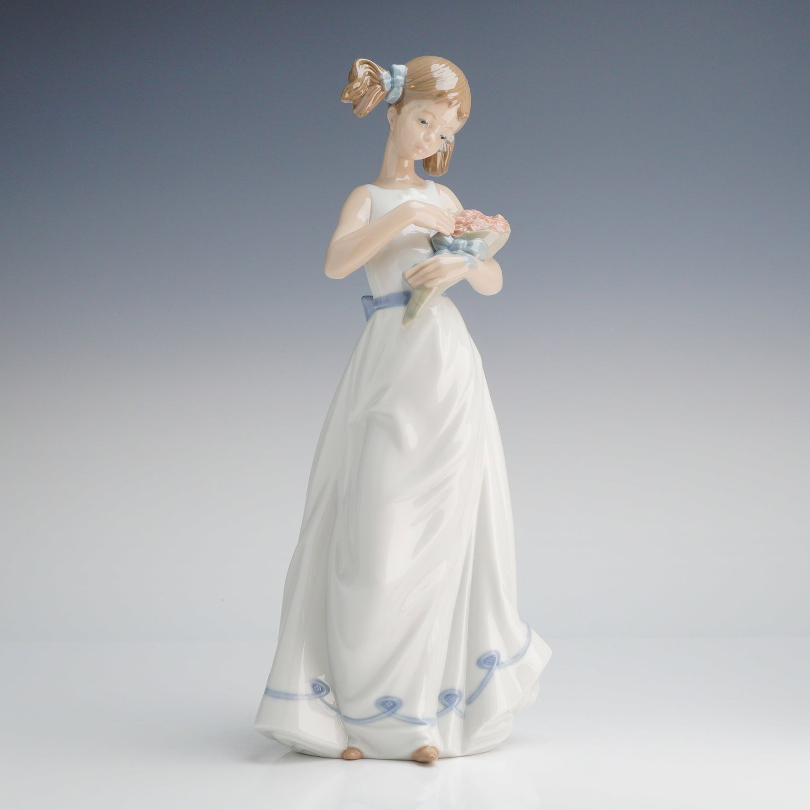 NAO by Lladro Porcelain Figurine, Girl with Holding a Bouquet: A NAO by Lladro glazed porcelain figurine of a girl with pigtails holding a bouquet. The figure is depicted in a standing pose, wearing a sleeveless white gown with a blue sash and a scrolled blue pat