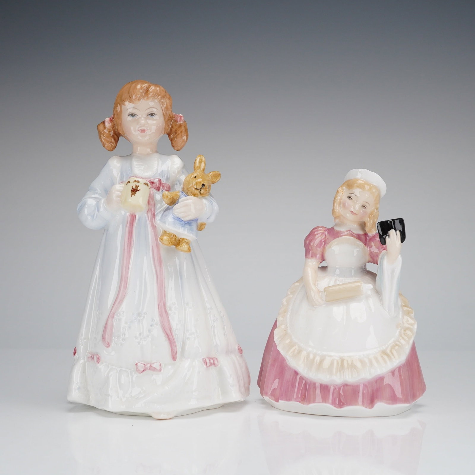 2pc Royal Doulton Figurines, Bunny's Bedtime HN 5231 and Cookie HN 2218: The first figurine, titled Bunny's Bedtime, depicts a young girl with pigtails dressed in a white nightgown accented with pink ribbons, holding a mug and a stuffed rabbit. The base of this figurine in