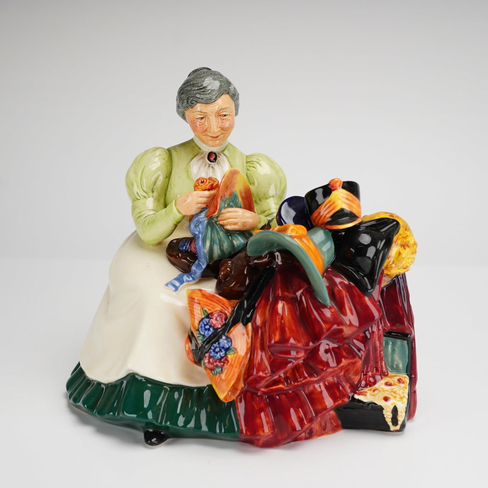 Royal Doulton Figurine The Wardrobe Mistress HN2145: A Royal Doulton glazed porcelain figurine titled The Wardrobe Mistress, model number HN 2145. The piece depicts an elderly woman with grey hair in a bun seated on a brown fringed stool. She is wearing
