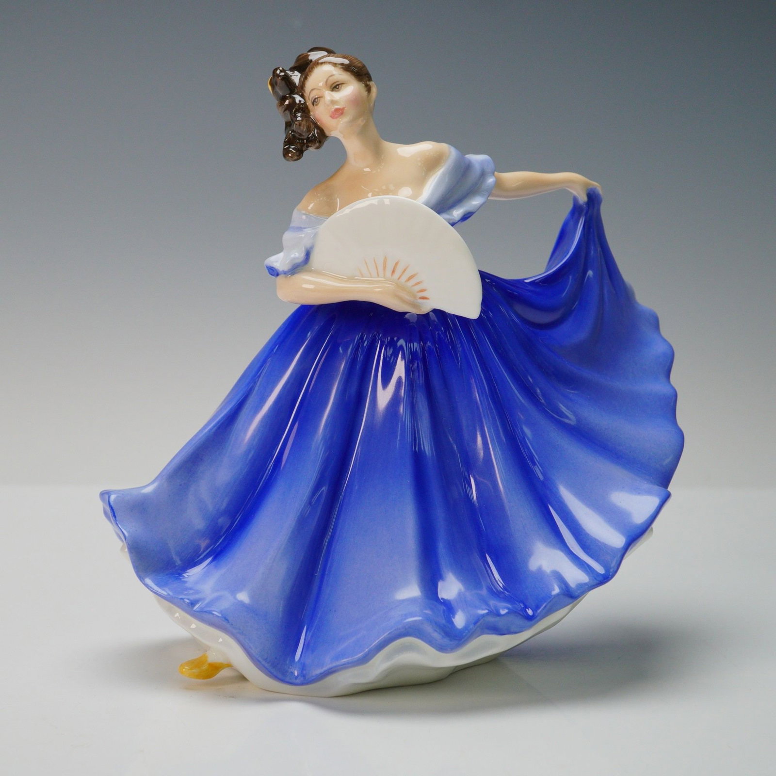 Royal Doulton Figurine, Elaine HN2791: A finely dressed lady in a royal blue gown holding a fan in one hand. Her brown hair styled in an elaborate updo adorned with a yellow ribbon. From the Peggy Davies Classic series. Royal Doulton backs