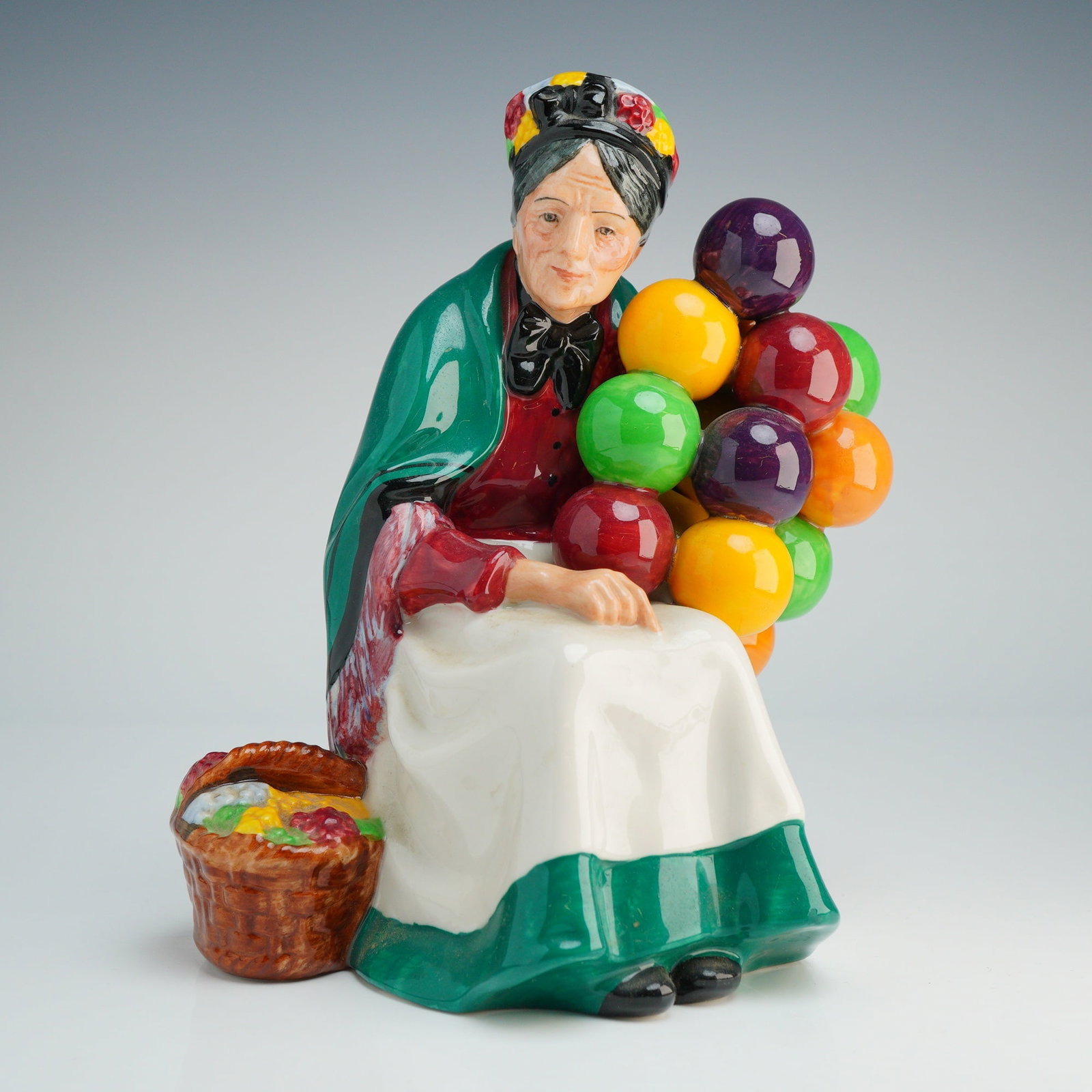 Royal Doulton Figurine, Old Balloon Seller HN1315: The figure depicts an elderly woman seated on a brown crate, wearing a red bodice, a green shawl with a fringed border, and a white apron with a green lower band. She holds a cluster of spherical ball