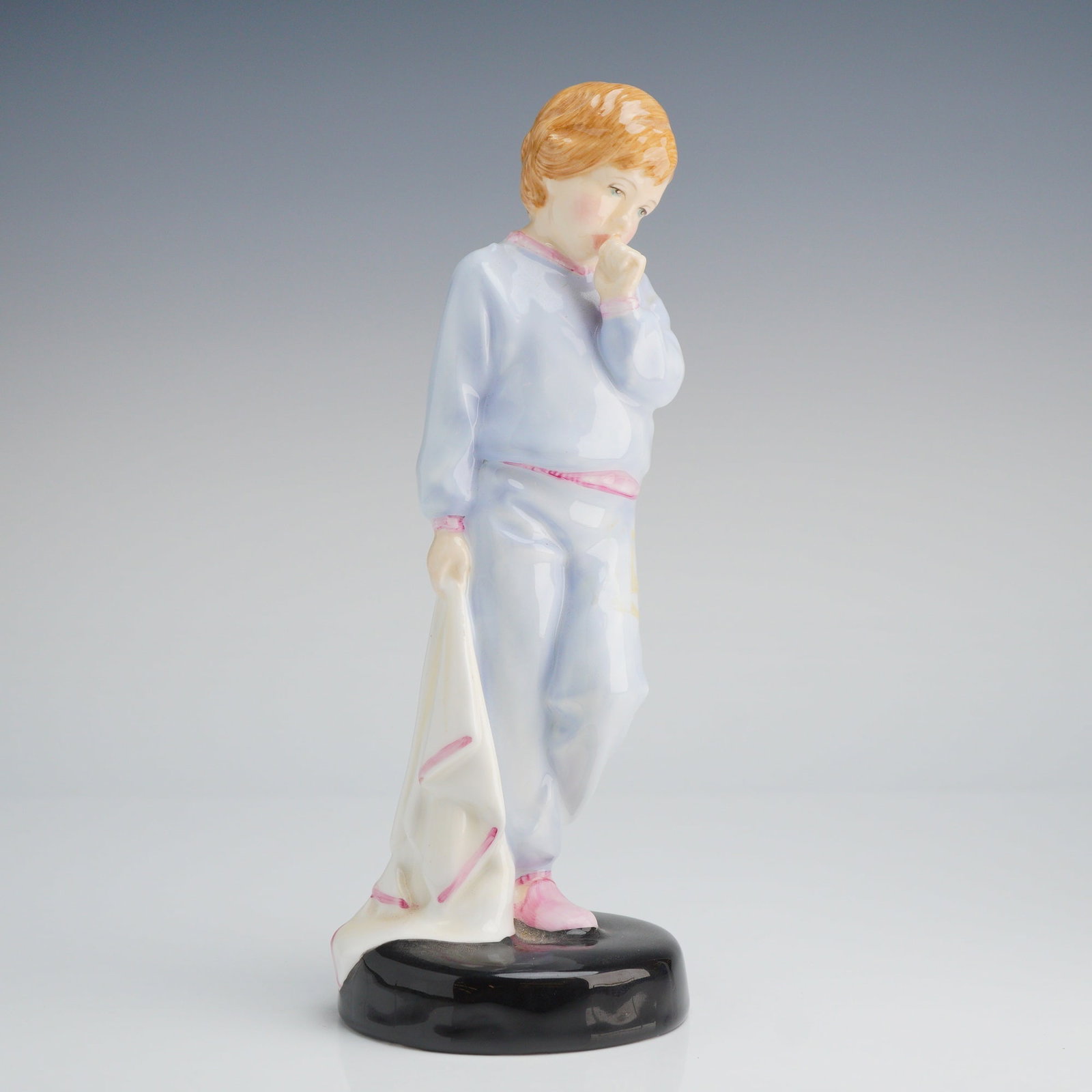 Royal Doulton Figurine, Sleepy Darling HN2953: The piece depicts a young child with reddish-blonde hair, dressed in light blue pajamas accented with pink trim, standing on a glossy black circular base while holding a white cloth or blanket. This f